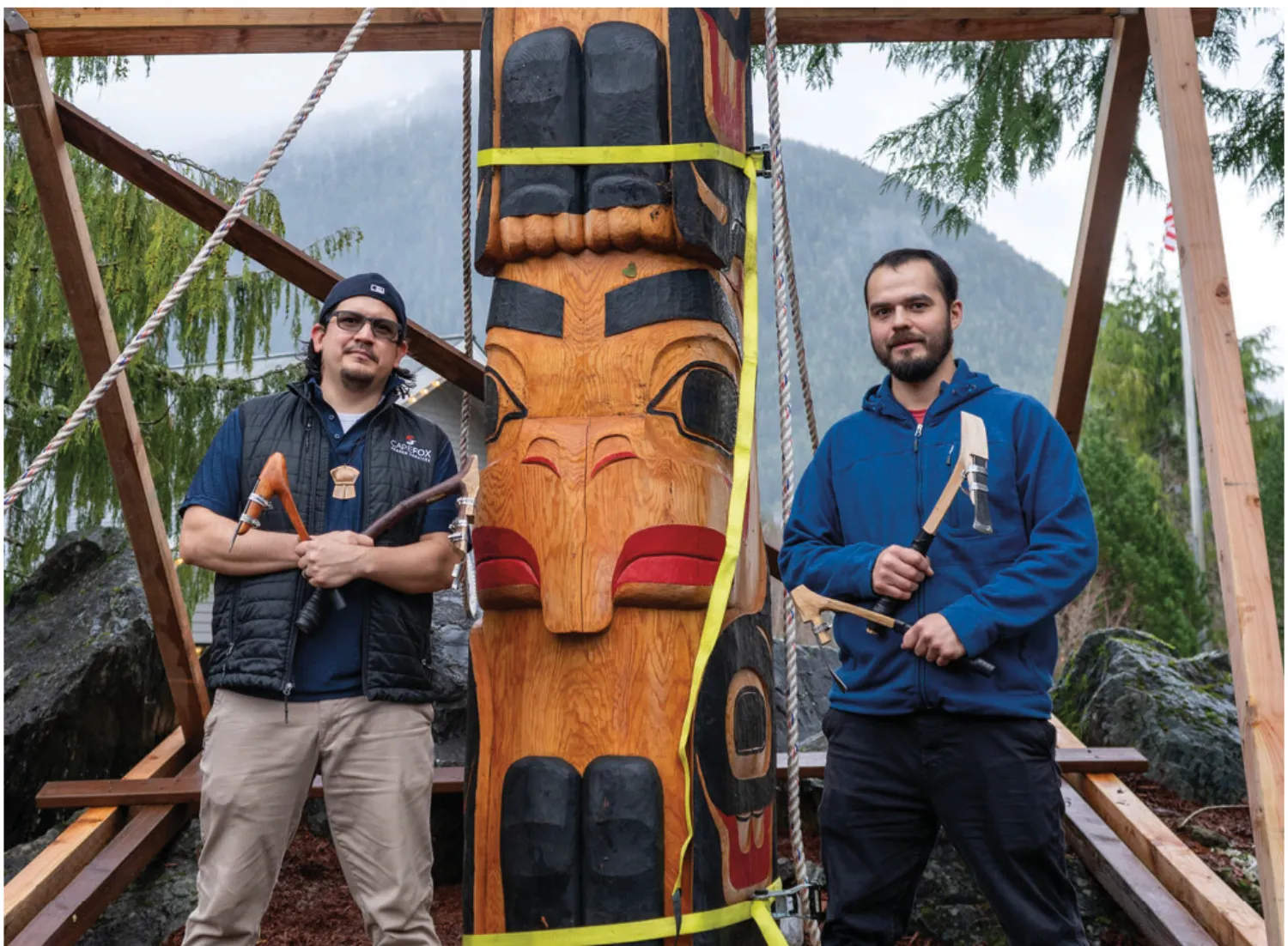 Two men stand in front of a totem pole they are carving, holding traditional woodworking tools, with trees and mountains in the background.