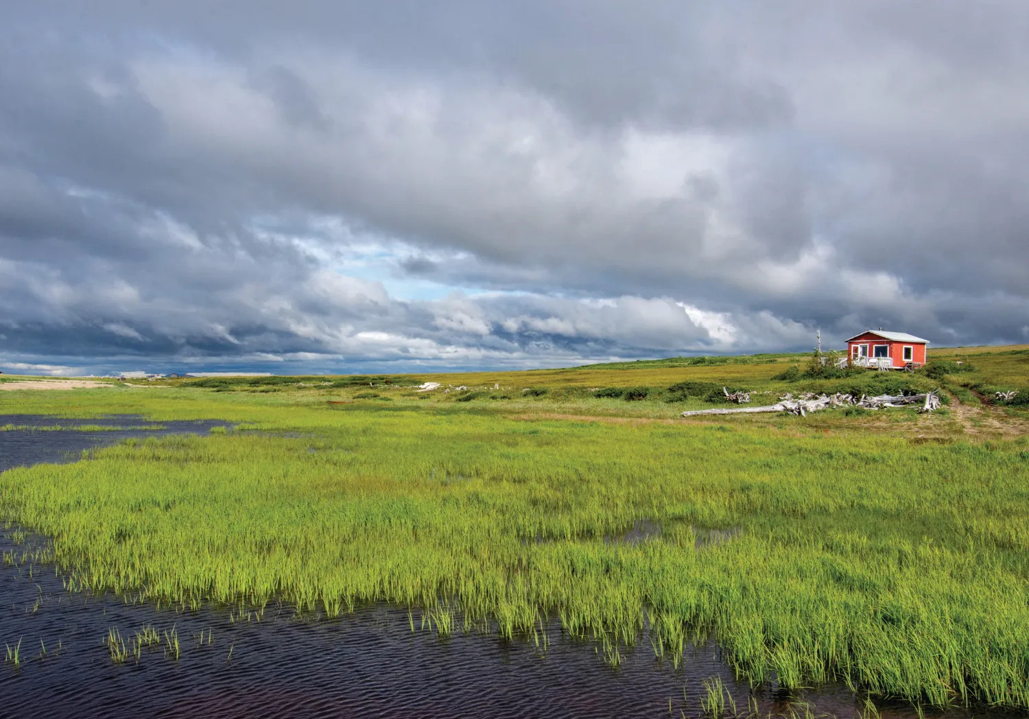 A remote landscape with green fields and a red cabin under a cloudy sky, showcasing the vast Alaskan wilderness.