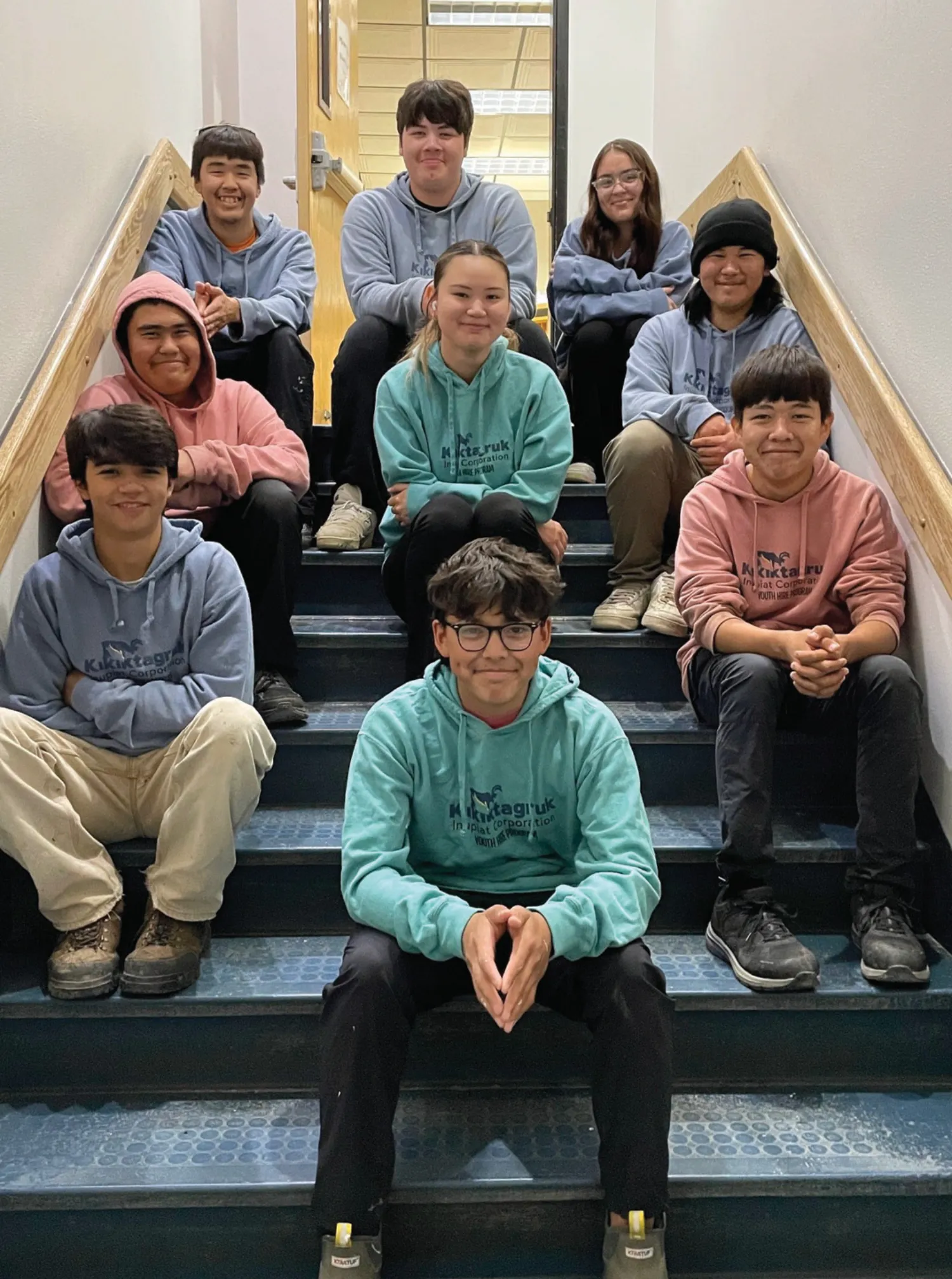 A group of youth seated on stairs, each wearing hoodies with logos, smiling at the camera indoors.