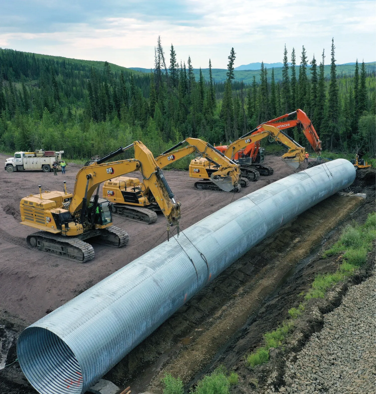 Multiple construction excavators working together to place a large metal culvert on a construction site surrounded by trees and hills.