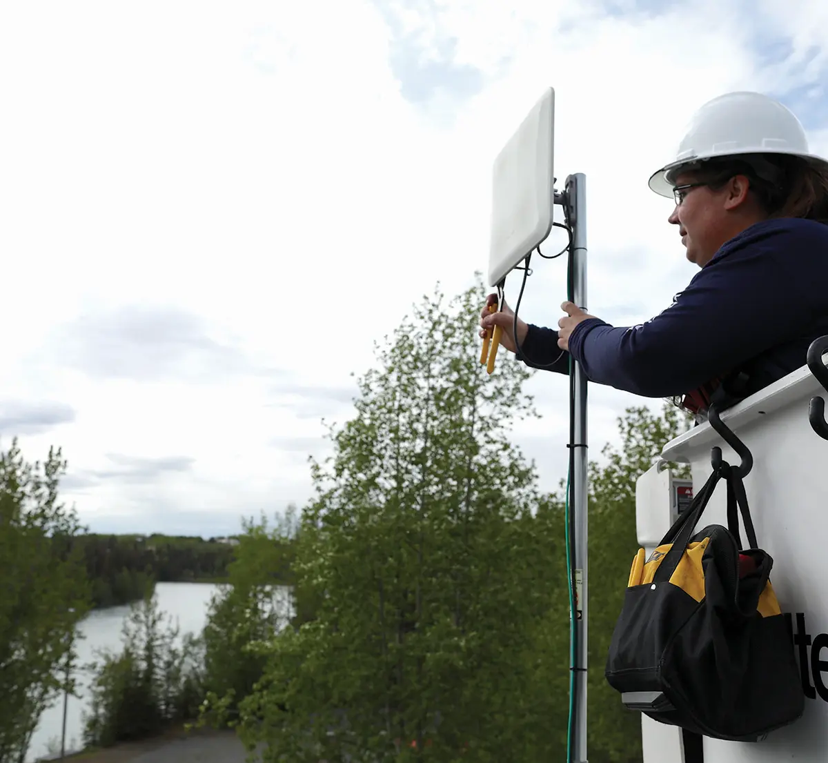 Portrait outdoor close-up photograph overcast view of a female Stantec technician employee worker with a white hardhat equipped and special safety glasses on who appears to be seen grasping a thin chrome broadband communication utility (possibly cellular communications) stick pole as she uses a yellow plier tool to cut a wire it looks like with her standing inside a white Stantec branded logo square shaped construction scissor lift or boom lift outside nearby some trees and a body of water plus there's a black/yellow branded DEWALT closed bag of tools it seems like hanging on one of the black hooks attached to the square shaped construction scissor lift or boom lift