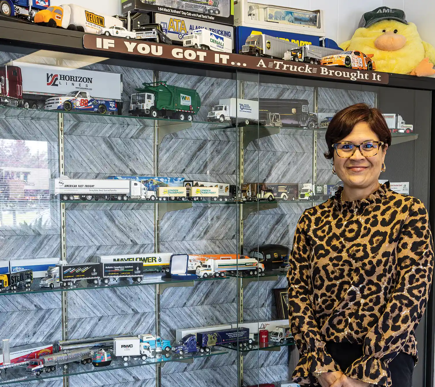 Jamie Benson wearing a leopard print top while posing next to a display of mini semi-trucks on shelves 