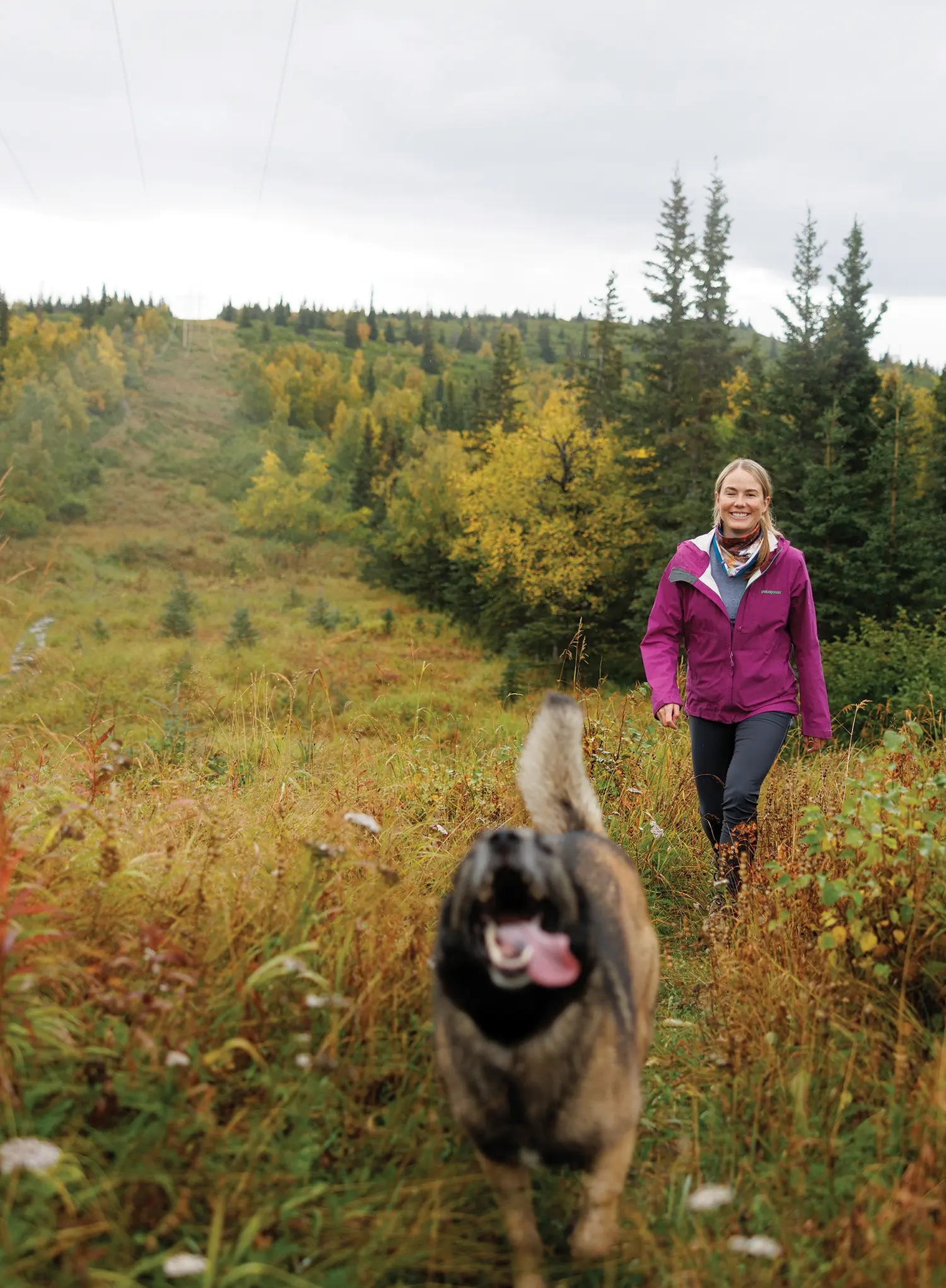 Portrait outdoor photograph close-up view of Jen Loofbourrow smiling in a dark magenta Patagonia branded windbreaker jacket, a bandana neck accessory around her, and black pants walking through the grassy meadows terrain as there's a dog sticking its tongue out in front of her while the dog is seen running away from her on a mostly cloudy overcast day