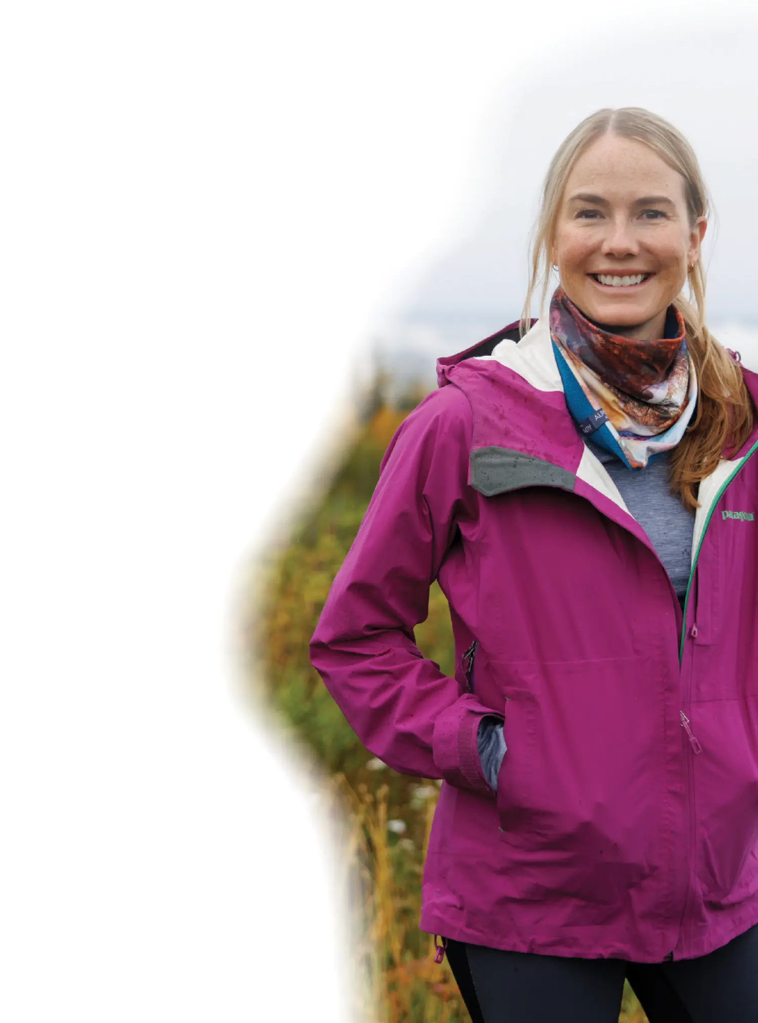Mostly white gradient background covering the portrait photograph close-up outdoor view of Jen Loofbourrow smiling in a dark magenta Patagonia branded windbreaker jacket, a bandana neck accessory around her, and black pants as she has her hands inside her pockets of the windbreaker jacket while she stands in a grassy meadows terrain on a mostly cloudy overcast day