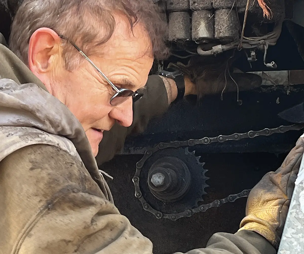 Portrait photograph of Bryce Wrigley in a beige colored jacket and tinted frame see through prescription glasses fixing internal components of the modern combine harvester machine with working gloves