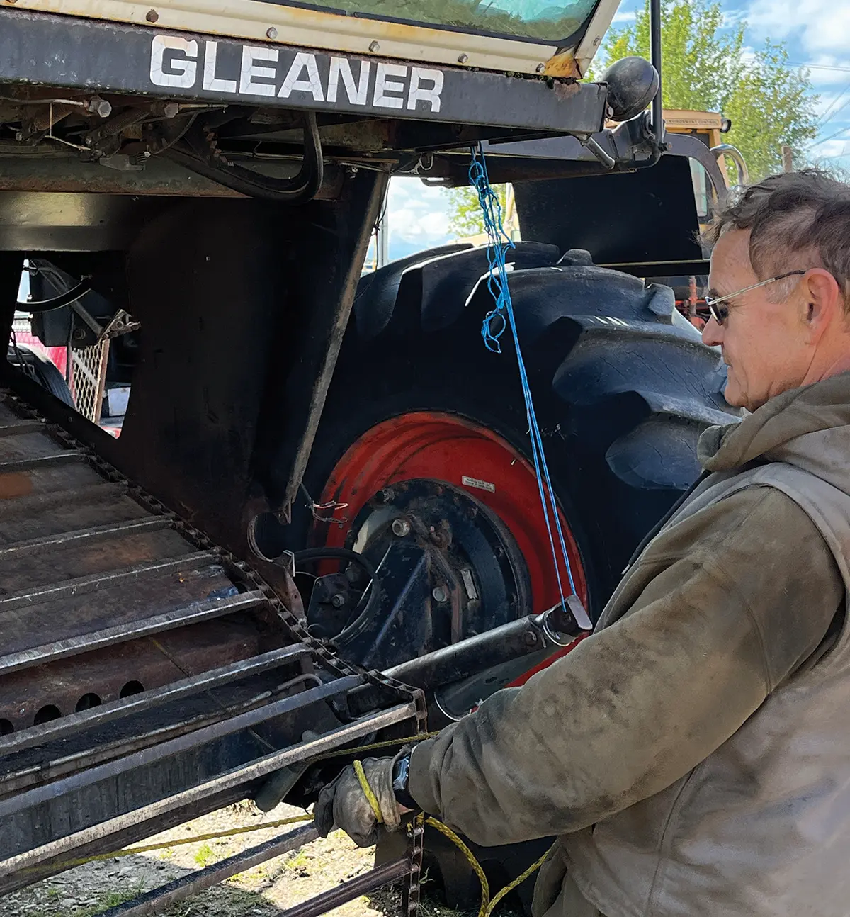 Portrait photograph of Bryce Wrigley in a beige colored jacket and tinted frame see through prescription glasses fixing the backside components of the modern combine harvester machine with working gloves