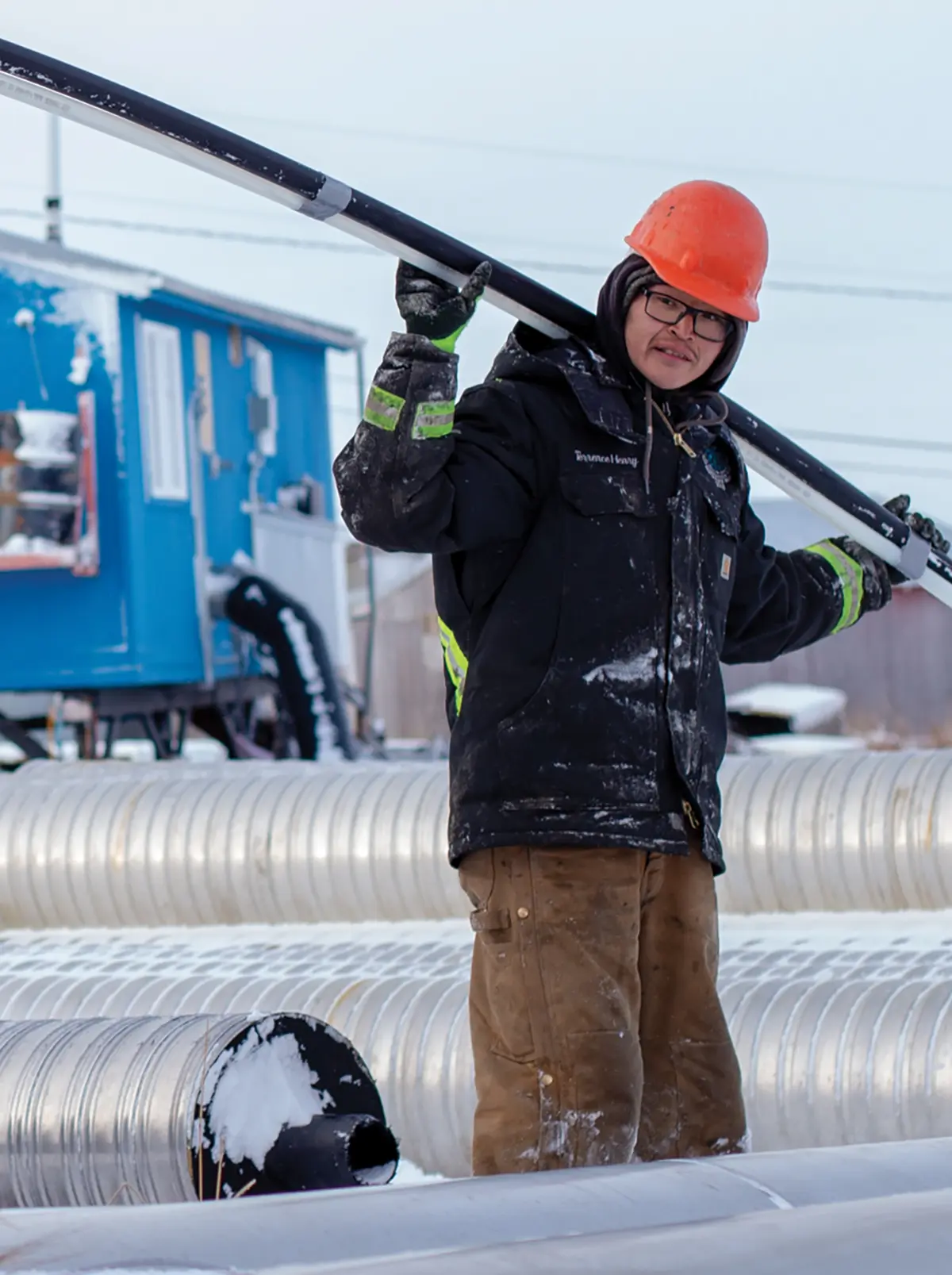 Portrait outdoor photograph view of a male Alaska Native Tribal Health Consortium (ANTHC) crew member in a black construction style puffy jacket, dark brown construction pants, and an orange hardhat holding a thin shaped black/white water/sewer pipeline around his shoulders as the crew completes the connection of one half of Akiachak to piped water and sewer for the first time on a cold, clear day