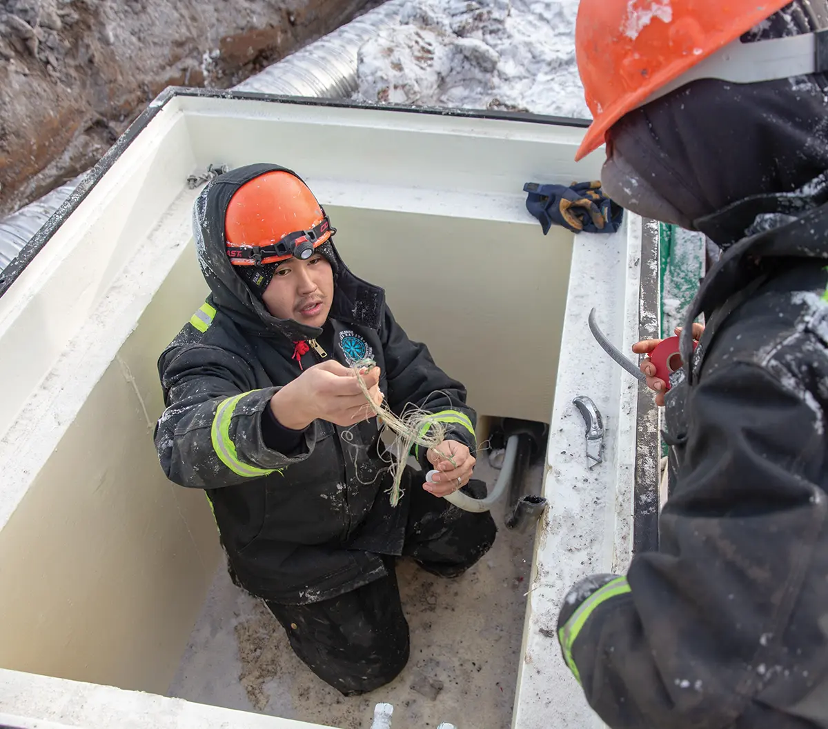 Portrait outdoor photograph view of a male Alaska Native Tribal Health Consortium (ANTHC) crew member and another ANTHC crew member watching upon this male ANTHC crew member as they complete a project in Eek installing a new point for raw water intake and a comprehensive piped water and sewer system as they are figuring out how to solve string style shaped wires that connect to the system; Both of these individuals are in a black construction style puffy jacket and an orange hardhat as this project provided the community and residents of Eek with in-home piped water and sewer for the first time