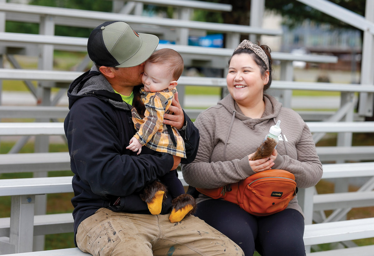 A family seated on outdoor bleachers with an adult kissing a baby and a smiling woman holding a bottle.