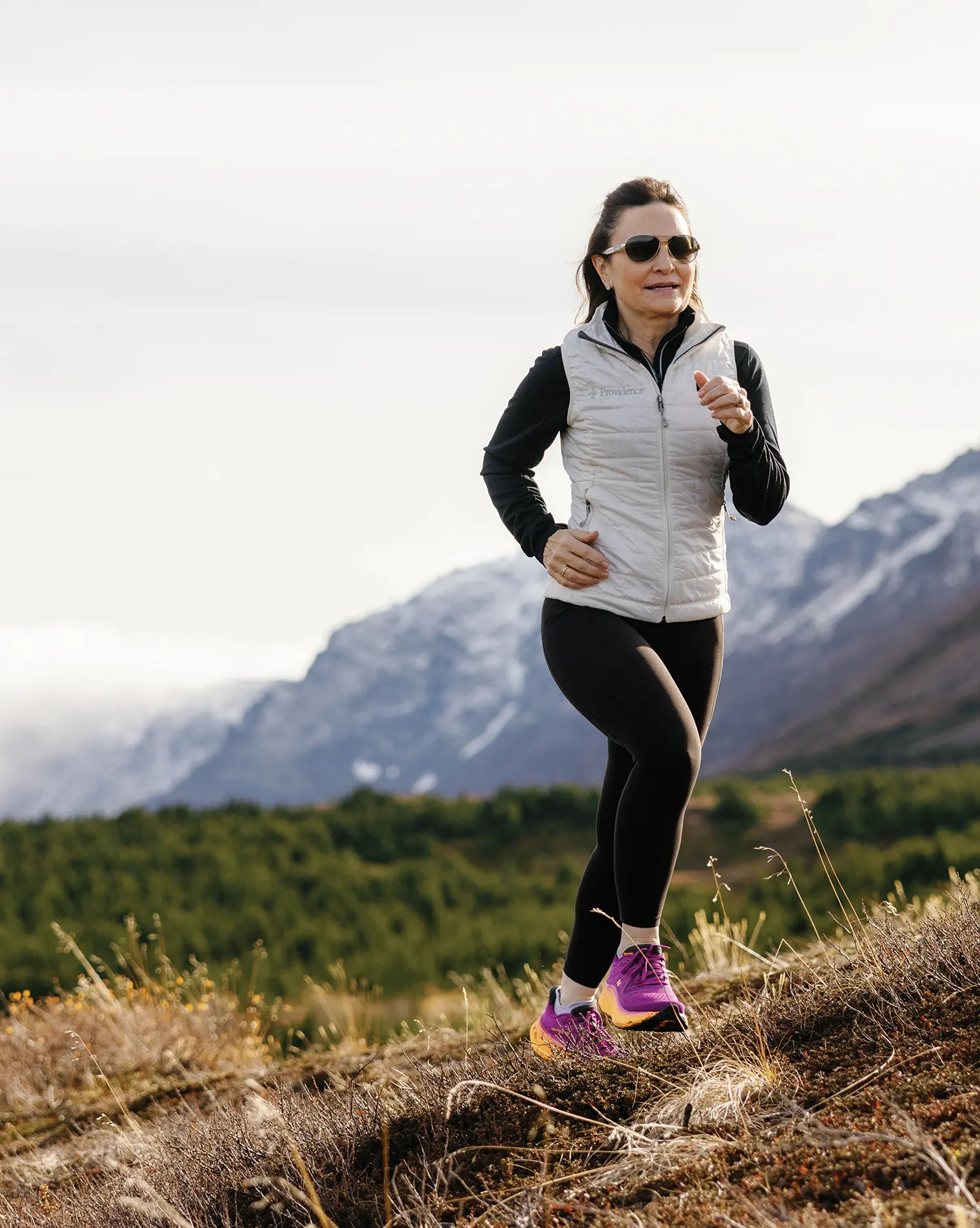 Portrait outdoor photograph close-up view of Ella Goss smiling in bronze/gold outer trim sunglasses, a light grey Patagonia branded vest, a black long-sleeve jacket underneath, black yoga sweatpants, and purple/orange New Balance fitness shoes as she is running up a hillside through the grassy meadows terrain on a mostly cloudy overcast day with a beautiful majestic mountain range plus many trees as the backdrop background behind her