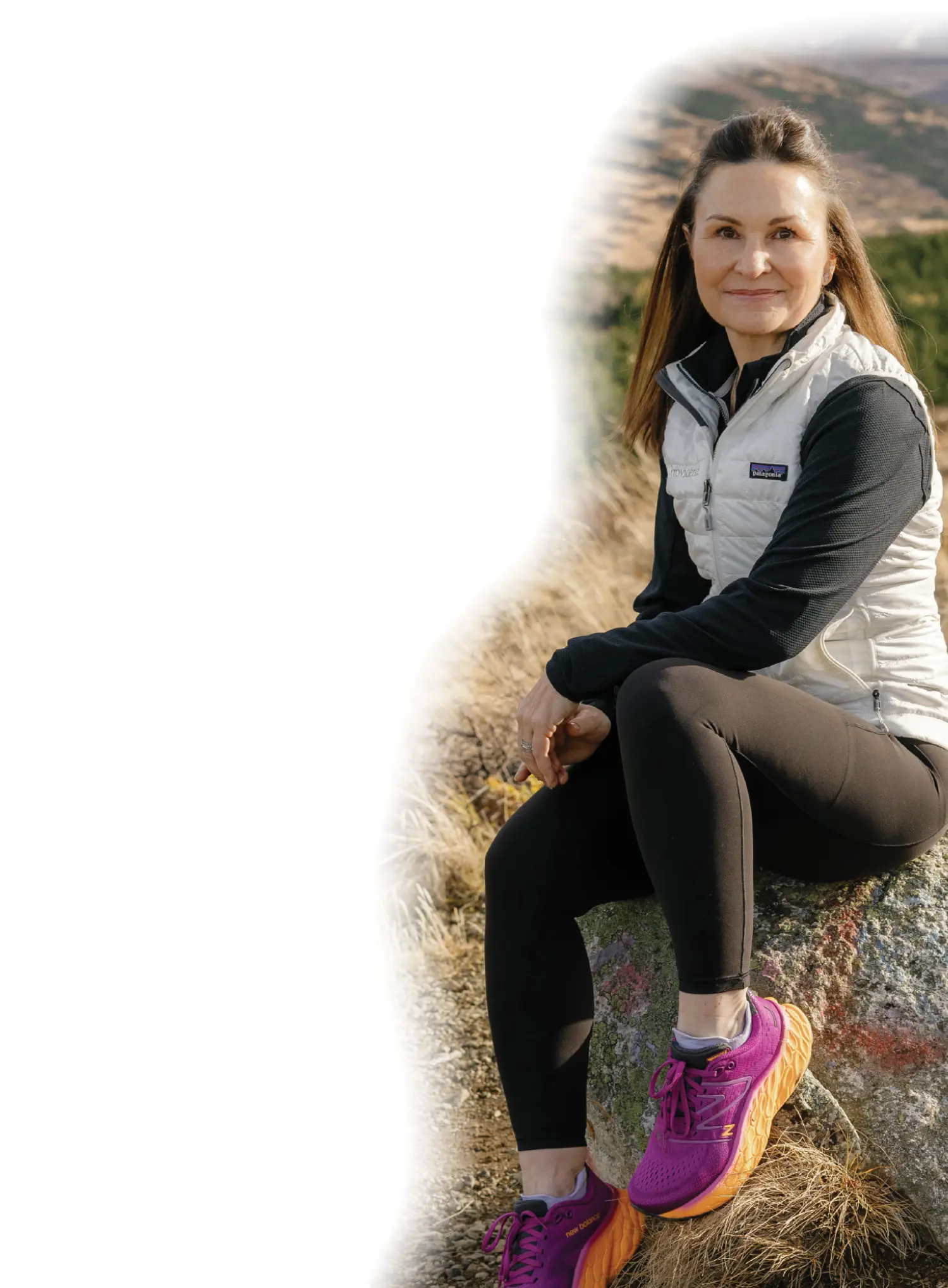 Mostly white gradient background covering the portrait photograph close-up outdoor view of Ella Goss grinning in a light grey Patagonia branded vest, a black long-sleeve jacket underneath, black yoga sweatpants, and purple/orange New Balance fitness shoes as she is sitting on a boulder rock on some hillside of the grassy meadows terrain on a mostly cloudy overcast day