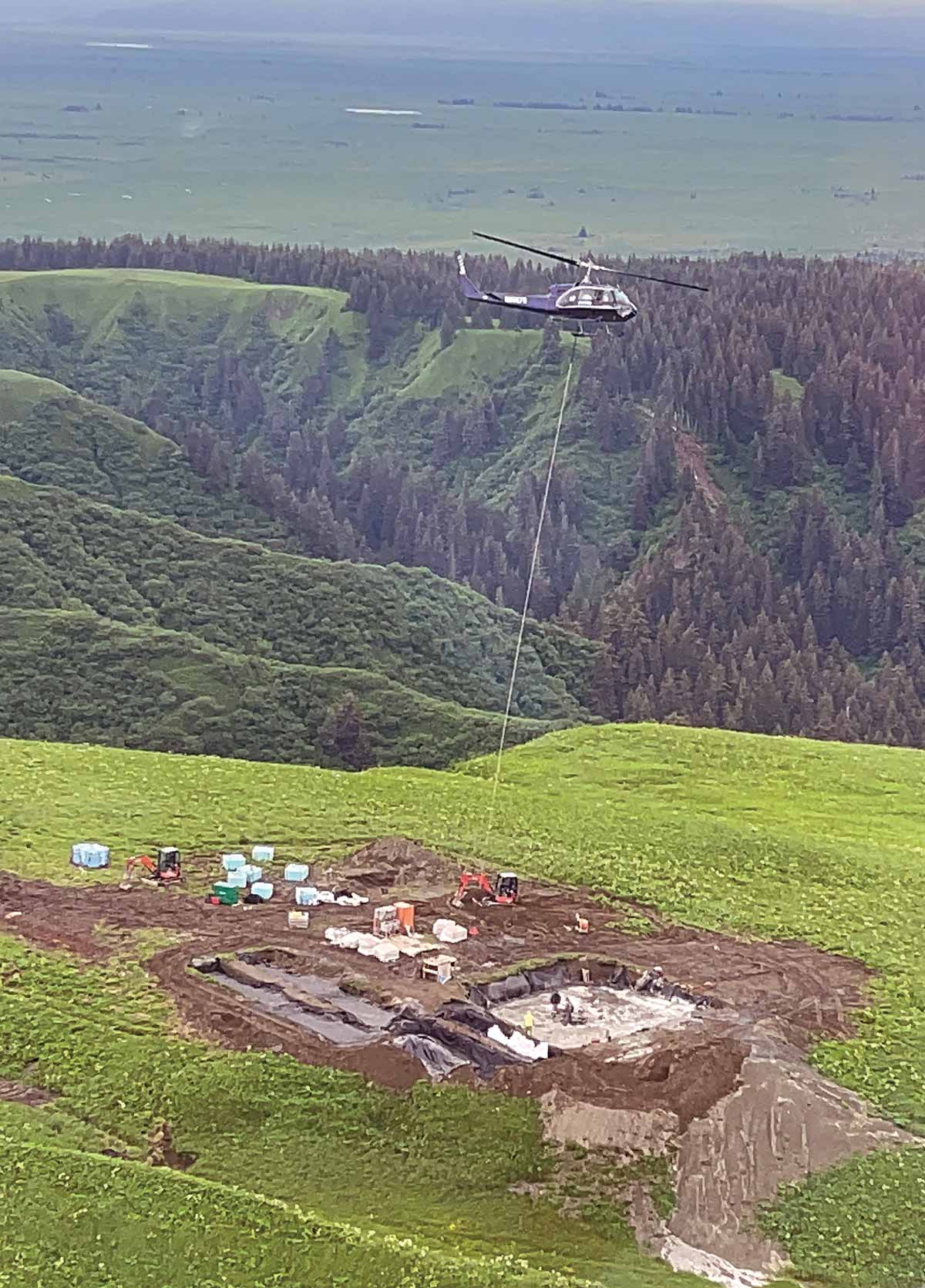A helicopter hovers over a remote, grassy construction zone carrying a large, white object suspended below