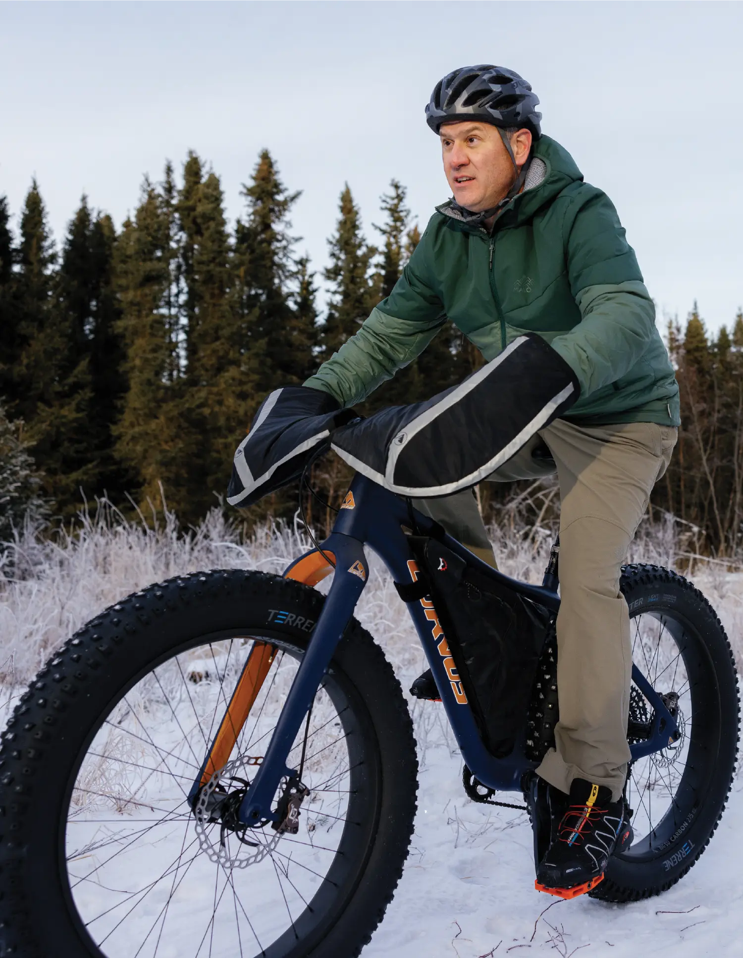 Portrait orientation outdoor photograph close-up view of Jason Metrokin riding in motion on top of a dark navy blue/orange/white outline colored mountain bicycle with big thick black wheels as he is looking up ahead in front of him at something off into the distance with big dark grey/black color combo covering hand cold snow mitts, a dark grey/black color combo camouflage bicycle helmet, dark forest/light forest green color combo thick puffy jacket, dark beige colored pants, and black/yellow/dark grey color combo mountain hiking shoes as he is situated on top of snowy covered ground with trees in the distance far behind him