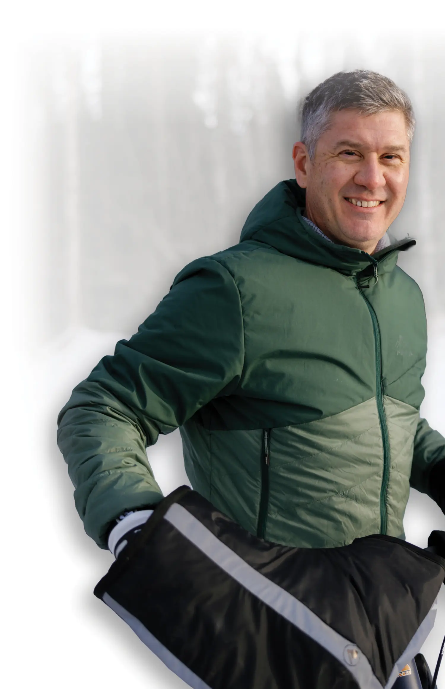 Mostly white to transparent gradient background covering the portrait orientation photograph close-up view of Jason Metrokin smiling with big dark grey/black color combo covering hand cold snow mitts and a dark forest/light forest green color combo thick puffy jacket as he is situated on top of his dark navy blue/orange/white outline colored mountain bicycle within snowy covered ground with trees in the distance far behind him