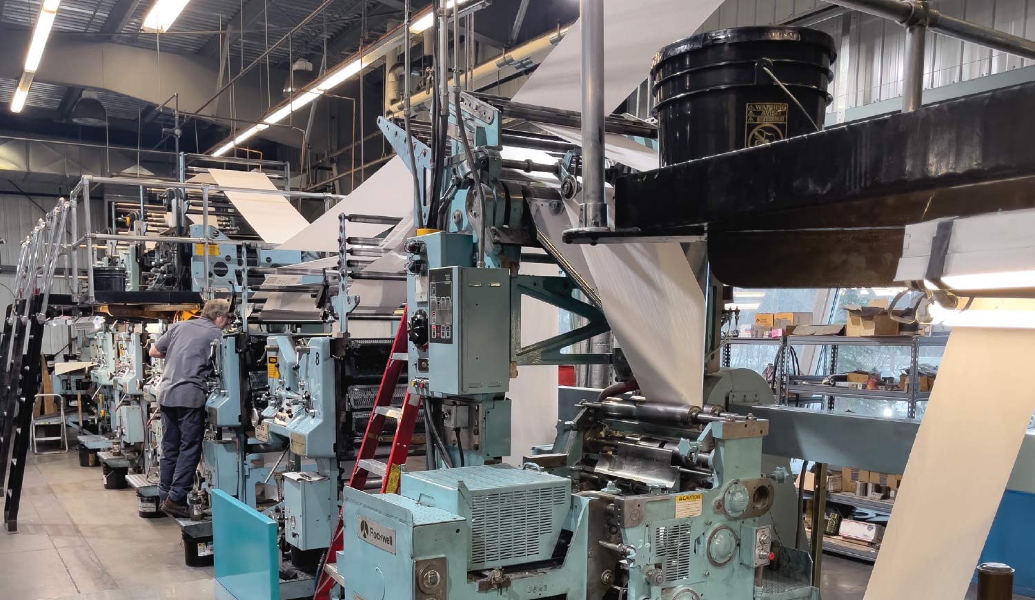 A pale blue and grey industrial printing press with a worker in a grey shirt