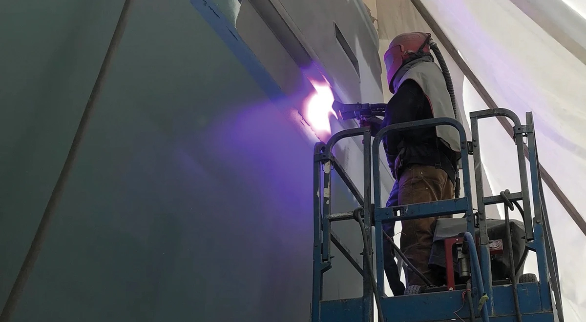 a person wearing a welding helmet and standing atop a gated lift applies thermal spray metalizing to a large structure