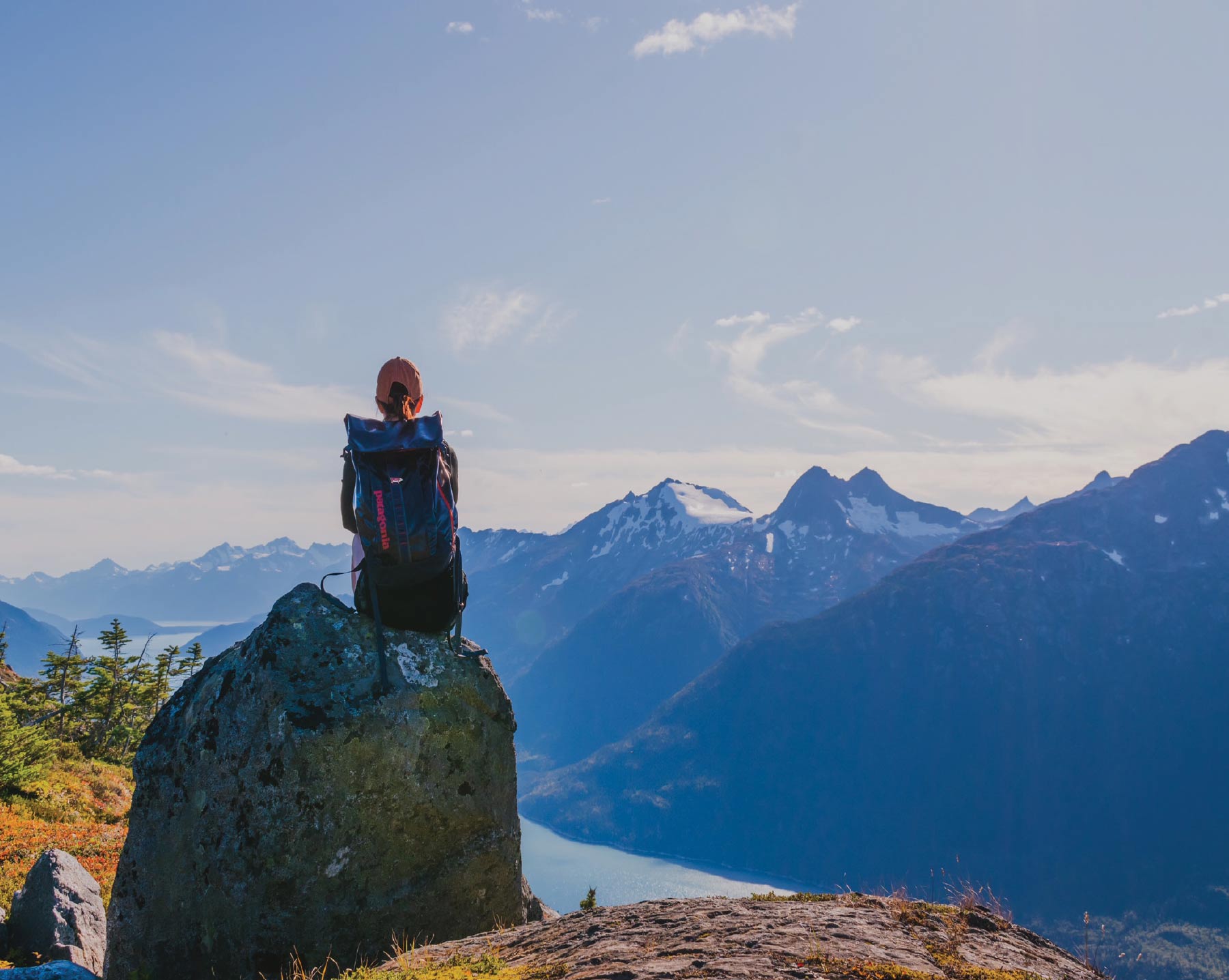 A person wearing a dark jacket and a large blue and black backpack, is sitting on a large rock overlooking a vast mountainous landscape with a winding body of water below, under a partly cloudy blue sky.