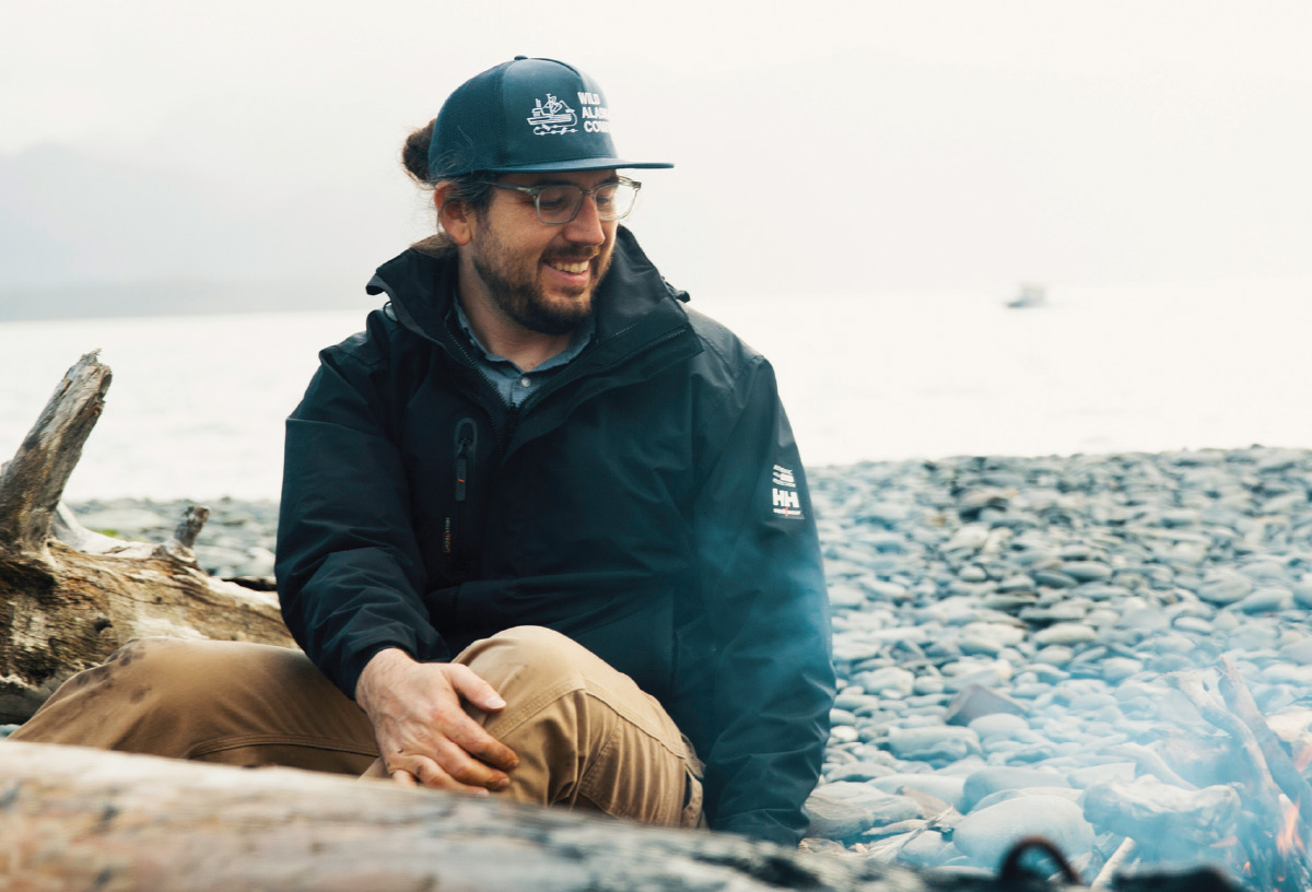 Arron Kallenberg wearing glasses, a dark jacket, a dark baseball cap, and tan pants is sitting on a piece of driftwood near a rocky shore and a body of water, looking down and smiling slightly.