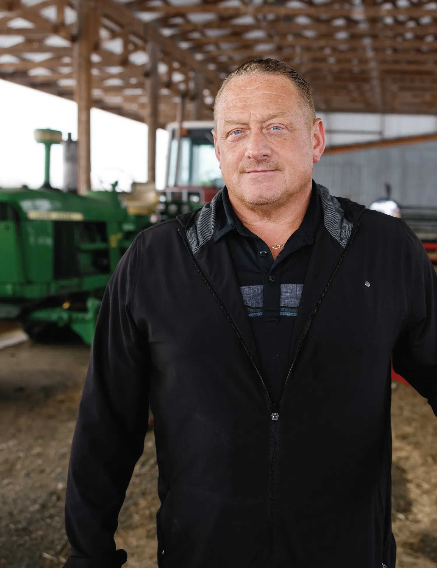 Portrait orientation outdoor close-up photograph view of Scott Hamilton grinning and standing for a pose as he is wearing a black jacket zipped closed halfway with a black polo top t-shirt underneath plus he has on a chrome necklace chain around him while he is inside a barn-like structure with farm equipment blurred out in the background behind him
