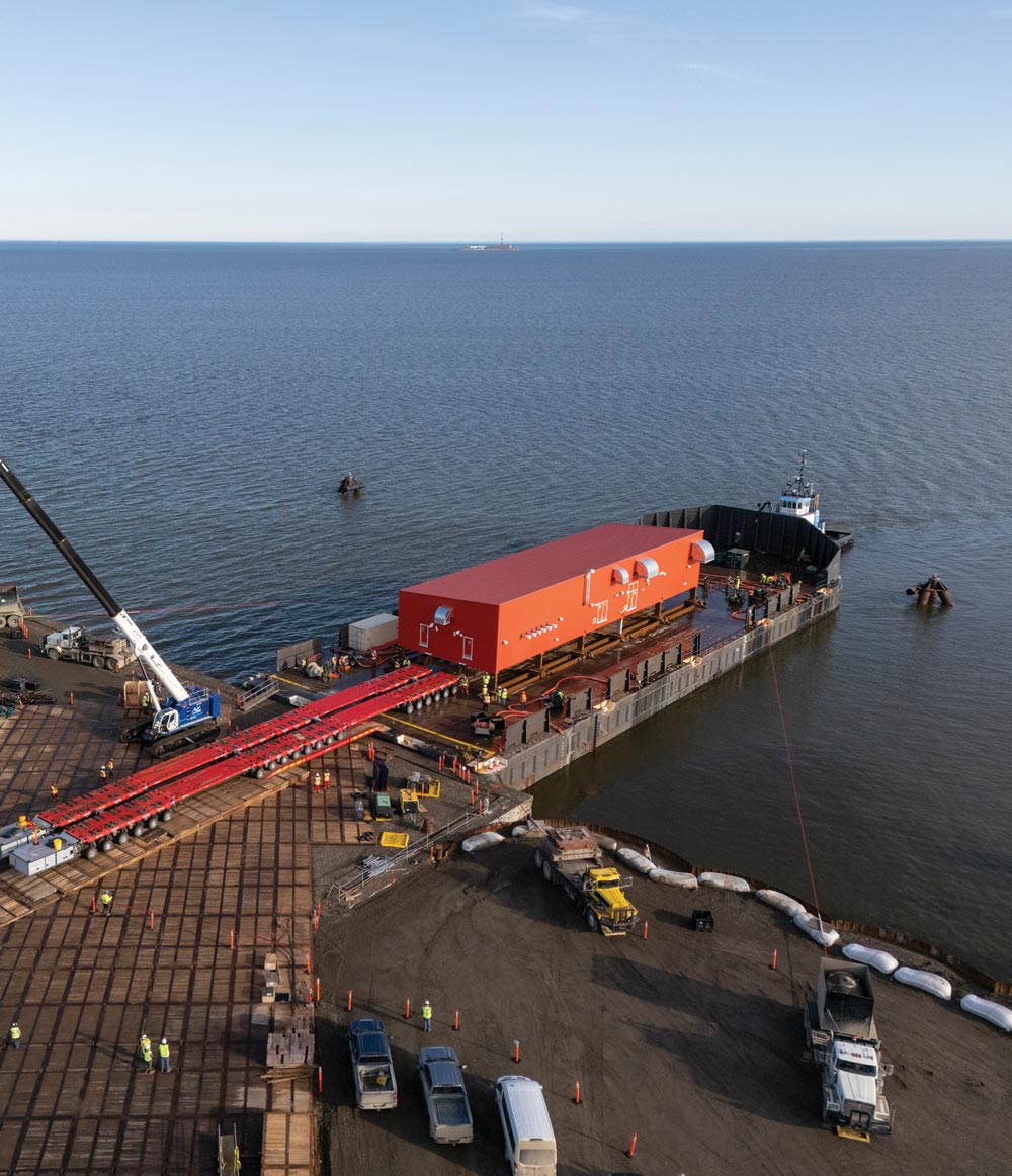 A large red structure being loaded onto a barge from a pier with construction vehicles and workers nearby.