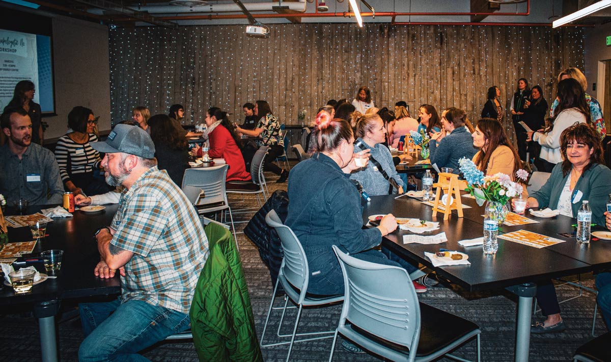 A social workshop with people seated at tables, engaging in conversations and dining in a dimly lit room with decorative lights on a wooden wall.