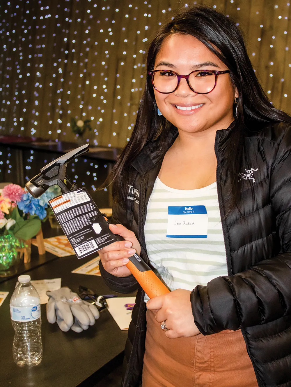 Person holding a hammer with a smile, standing in front of a table with various items.