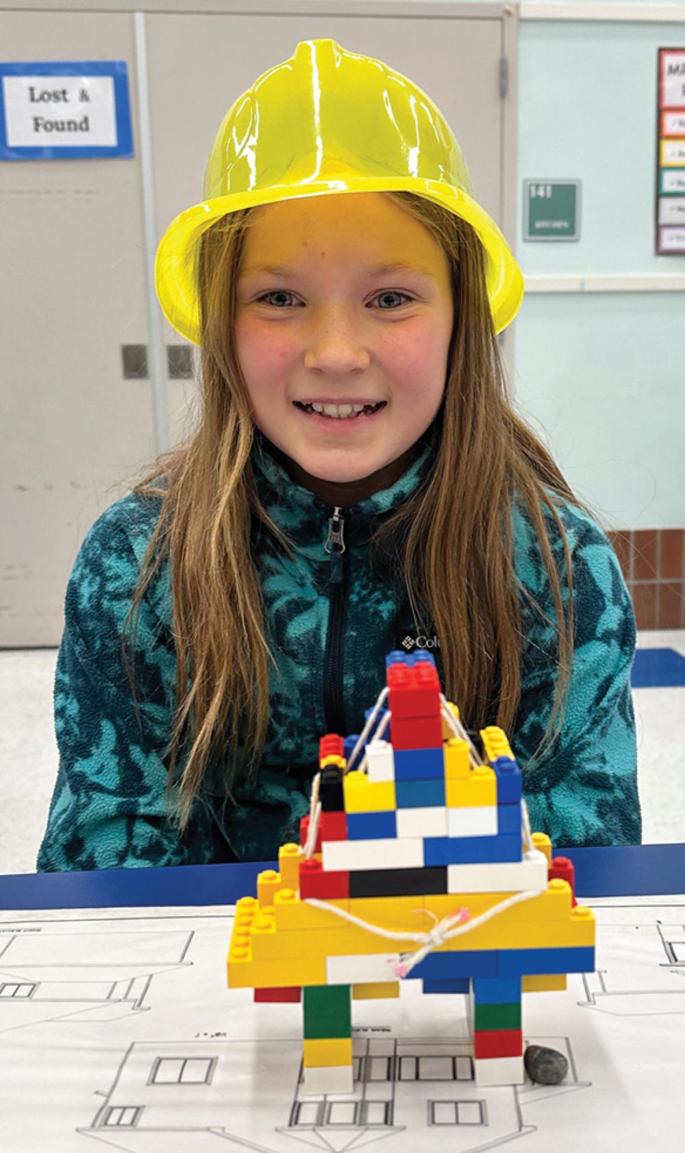 Young girl wearing a yellow construction helmet with a LEGO structure on the table in front of her.