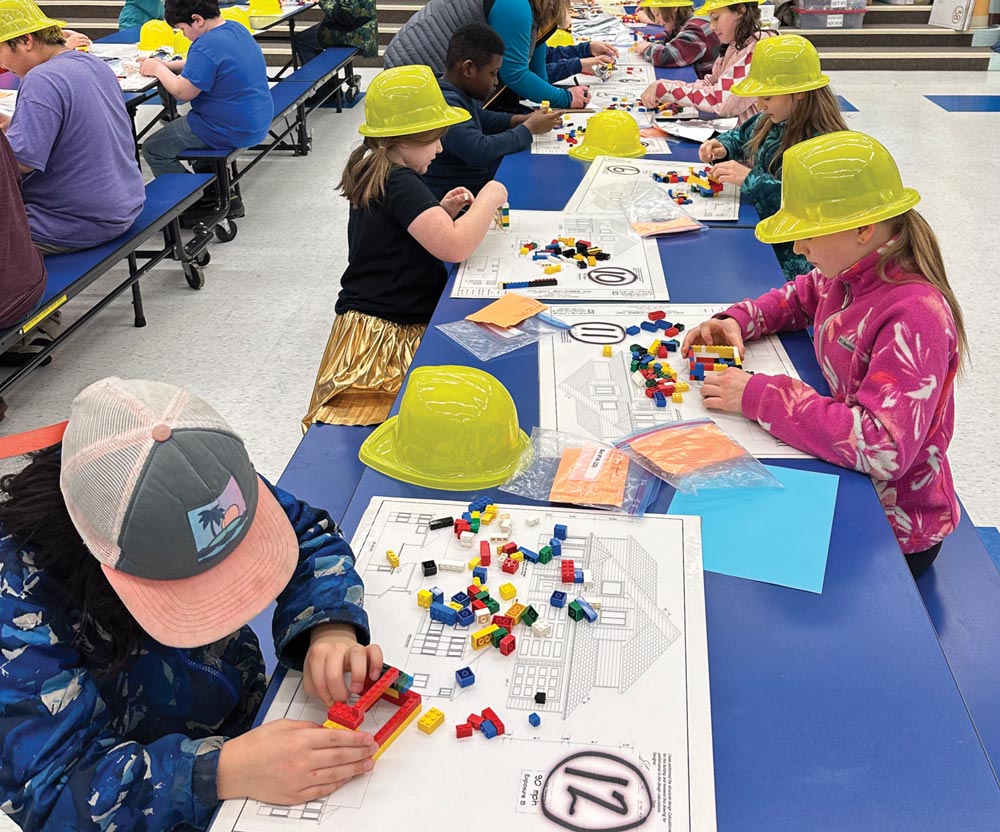 Children building LEGO models at tables with construction hats in a classroom.