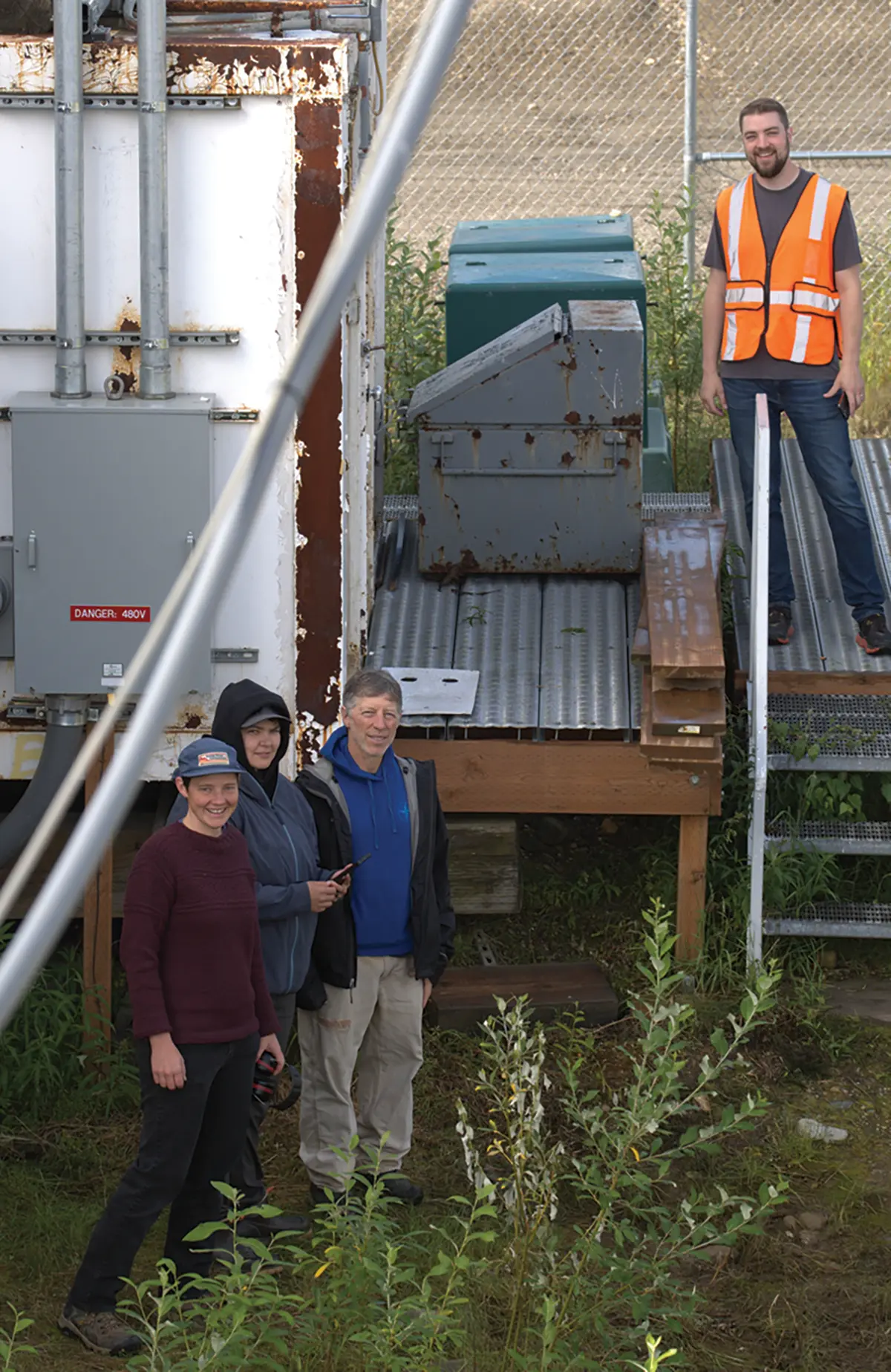 Portrait orientation outdoor photograph of four people standing and smiling outdoors nearby industrial equipment and a chain-link fence as one individual out of this group is wearing a high-visibility orange safety construction vest with everyone situated in Shungnak, Alaska