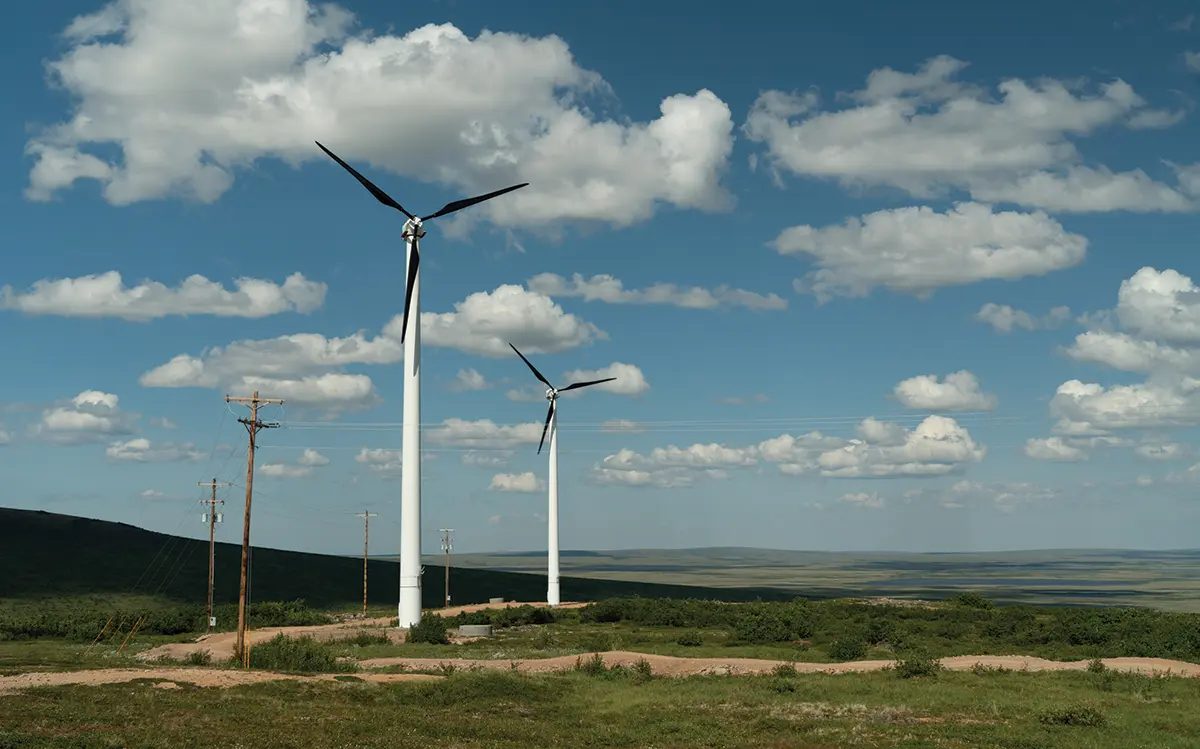 Portrait orientation outdoor photograph of two white Vestas V110-2.0 wind turbines with black blades against a blue clear sky containing scattered white clouds as the wind turbines are visibly standing tall situated in a rural landscape area with rolling sprawling hills and power lines in Buckland, Alaska