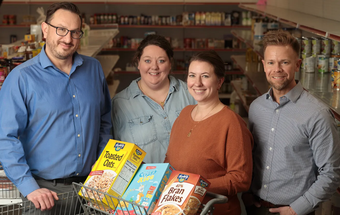 A group of four people standing in front of shelves with food items, smiling. They are holding various food boxes, including Toasted Oats, Crispy Rice, and Bran Flakes, with a cart in front of them.