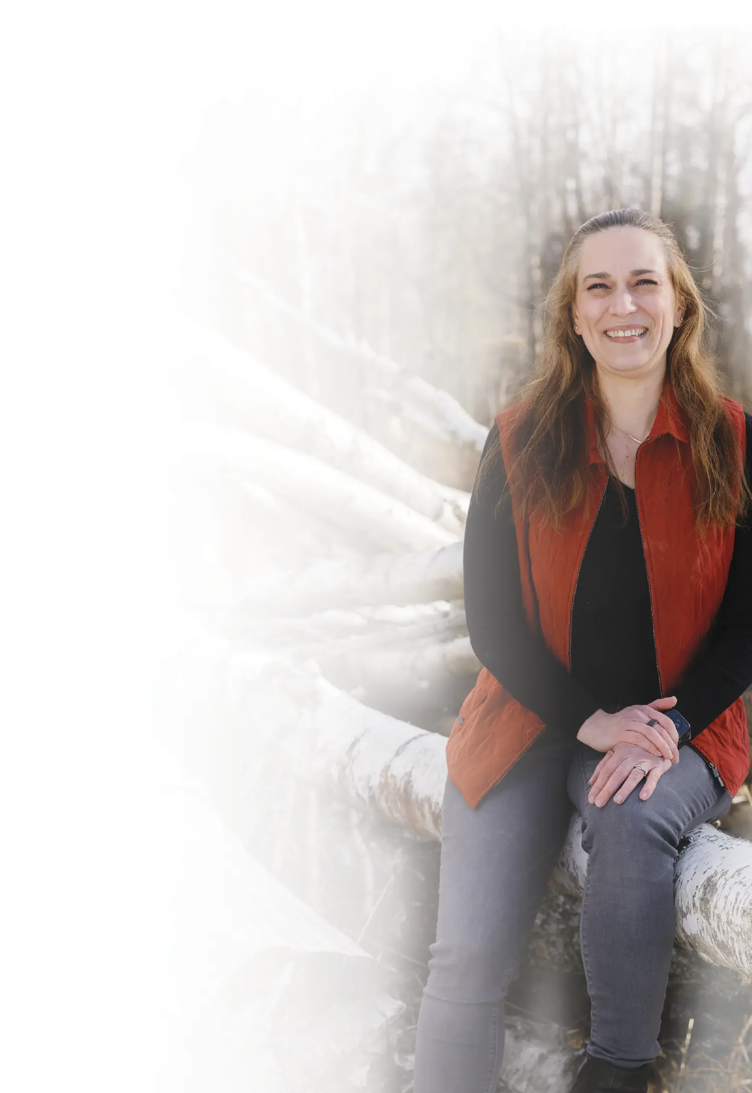 Mostly white to transparent gradient background covering the portrait orientation photograph close-up view of Christine Hopkins smiling and seated down for a pose in long brown hair as she is wearing a red quilted vest over a black long-sleeved sweatshirt and dark gray jeans with her situated on top of a light-colored log outside in nature; The background is softly blurred out with trees and a hint of sky