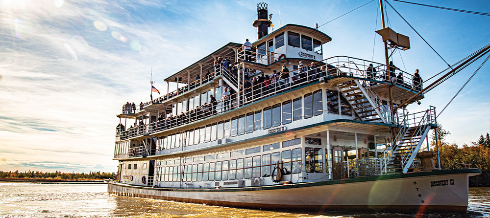 A large, multi-deck riverboat, "Discovery III," is on a river with many passengers visible.