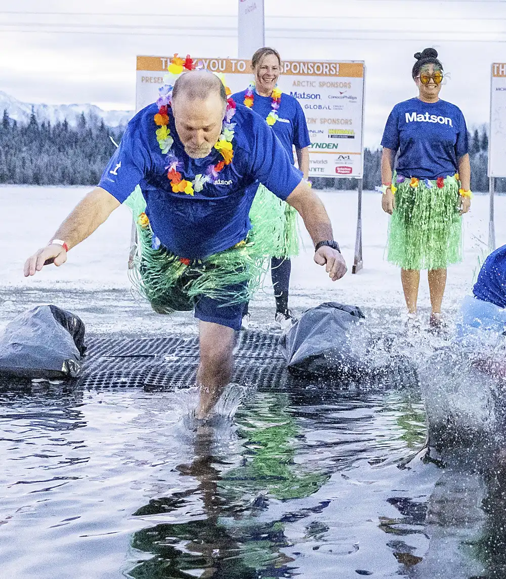 A man wearing a lei and hula skirt splashes into icy water, with two women in similar outfits standing behind him.