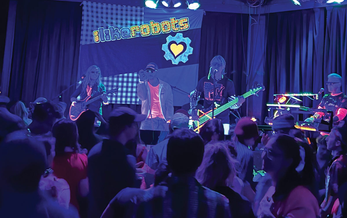 the band, “I Like Robots,” performs on stage under blue and purple lights for a crowd, with a banner featuring their name and a heart icon