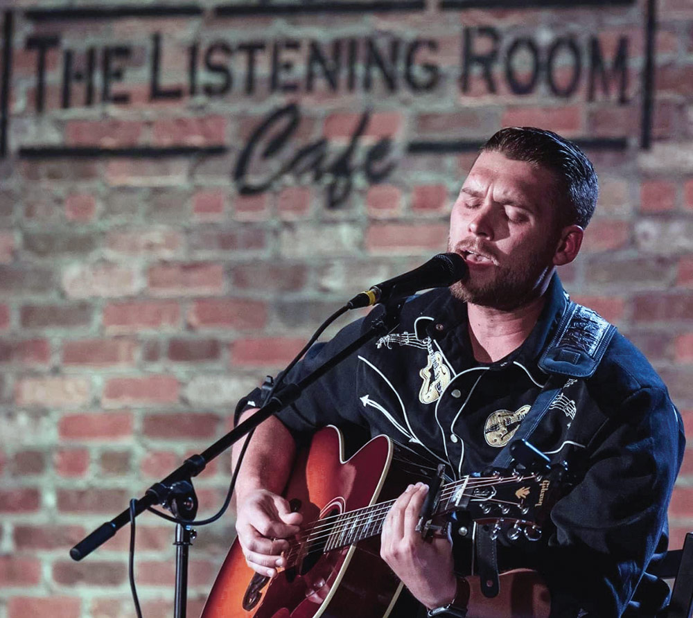 Schaefer Mueller sings into a microphone while playing an acoustic guitar, in front of a brick wall with "THE LISTENING ROOM Cafe" written on it.