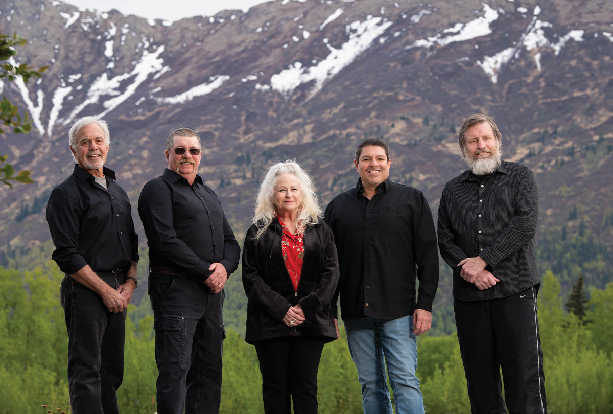 The Glacier Blues Band members, four men and one woman, stand in front of a snow-capped mountain range and green trees.