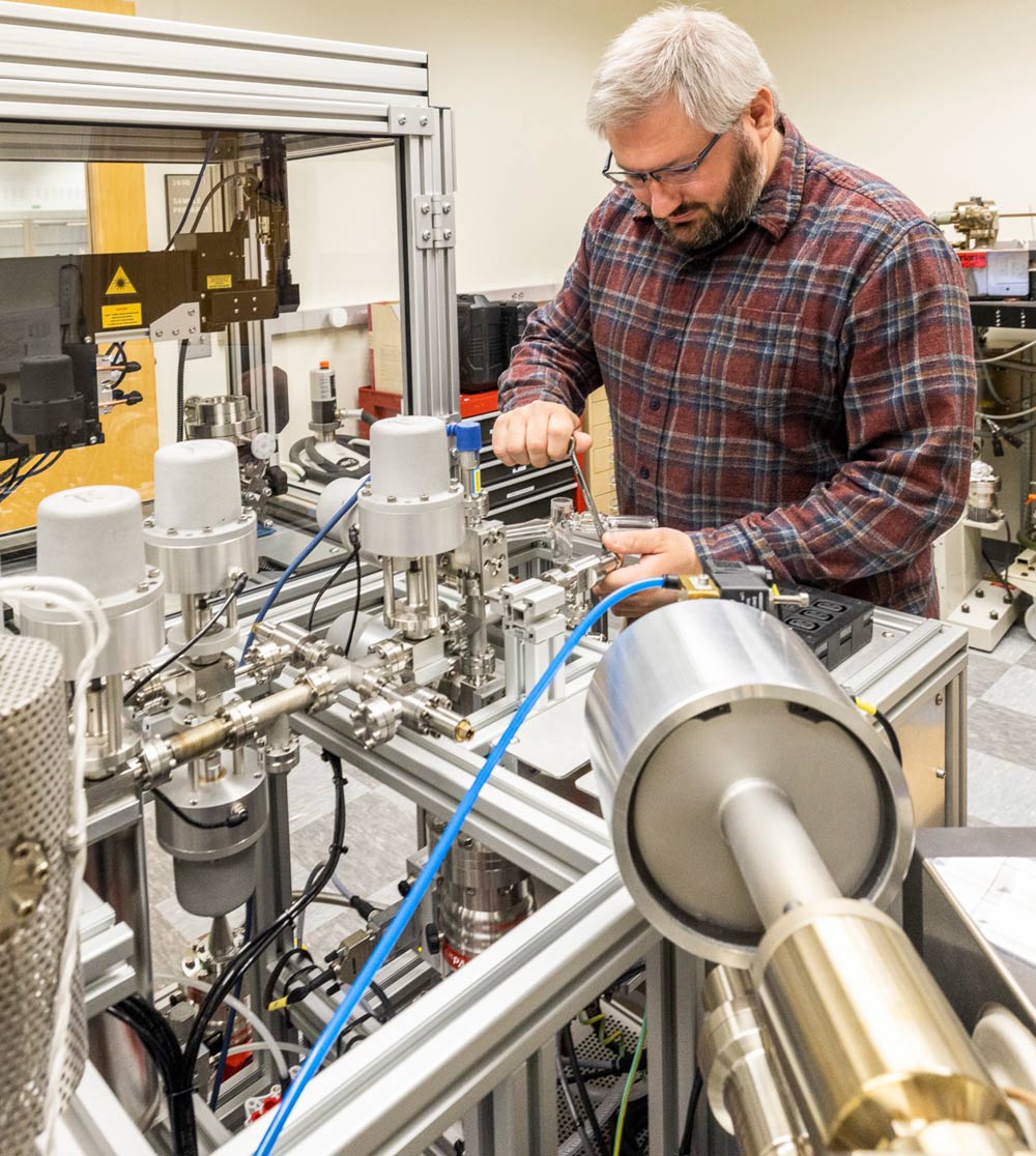 A man in a plaid shirt works on complex laboratory equipment involving metal cylinders and tubes.