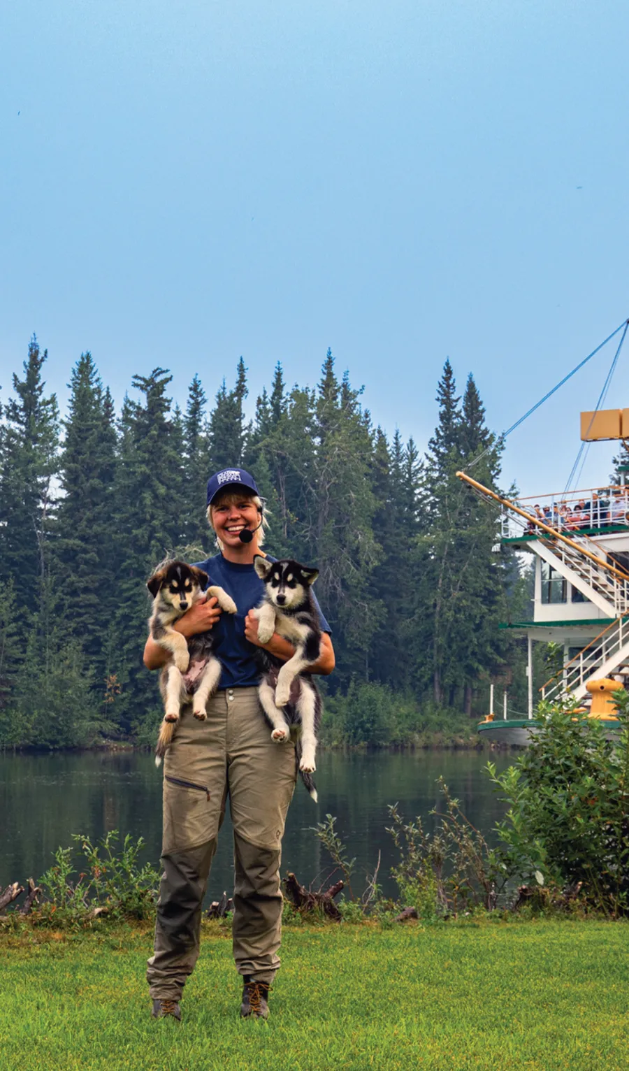 Susan Butcher holding two puppy sled dogs with the forest and a steamboat behind her