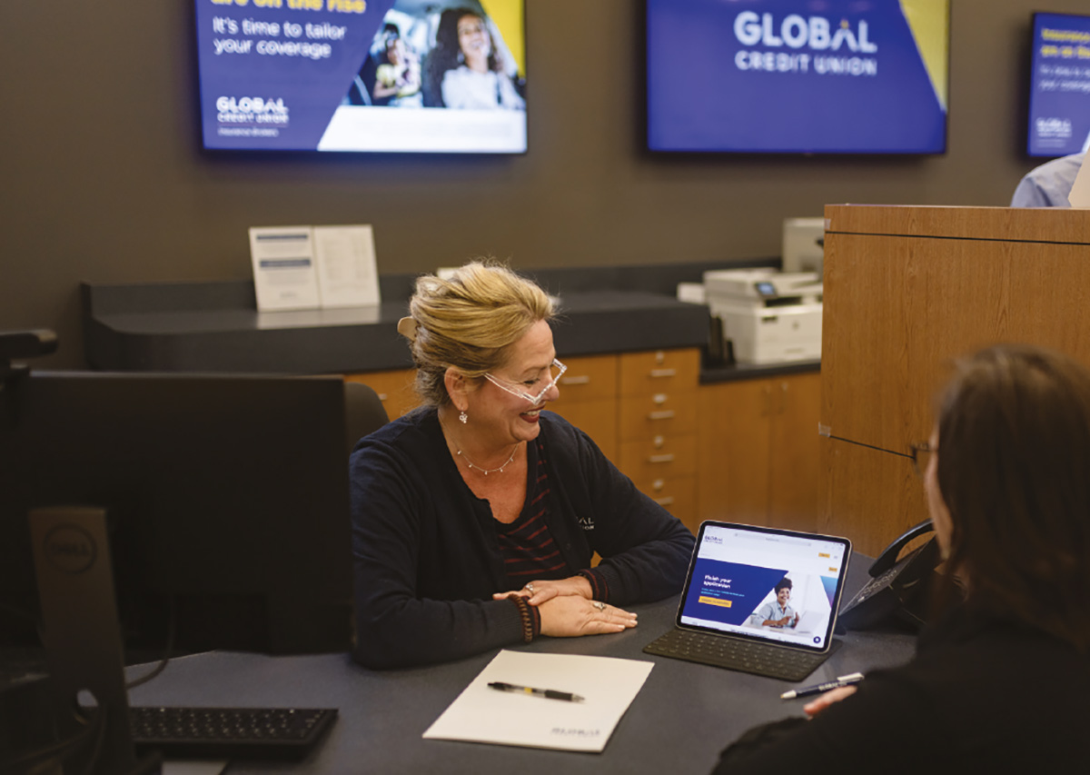 A female global credit union employee with blonde hair and glasses smiles while looking at a client sitting across a desk from her.