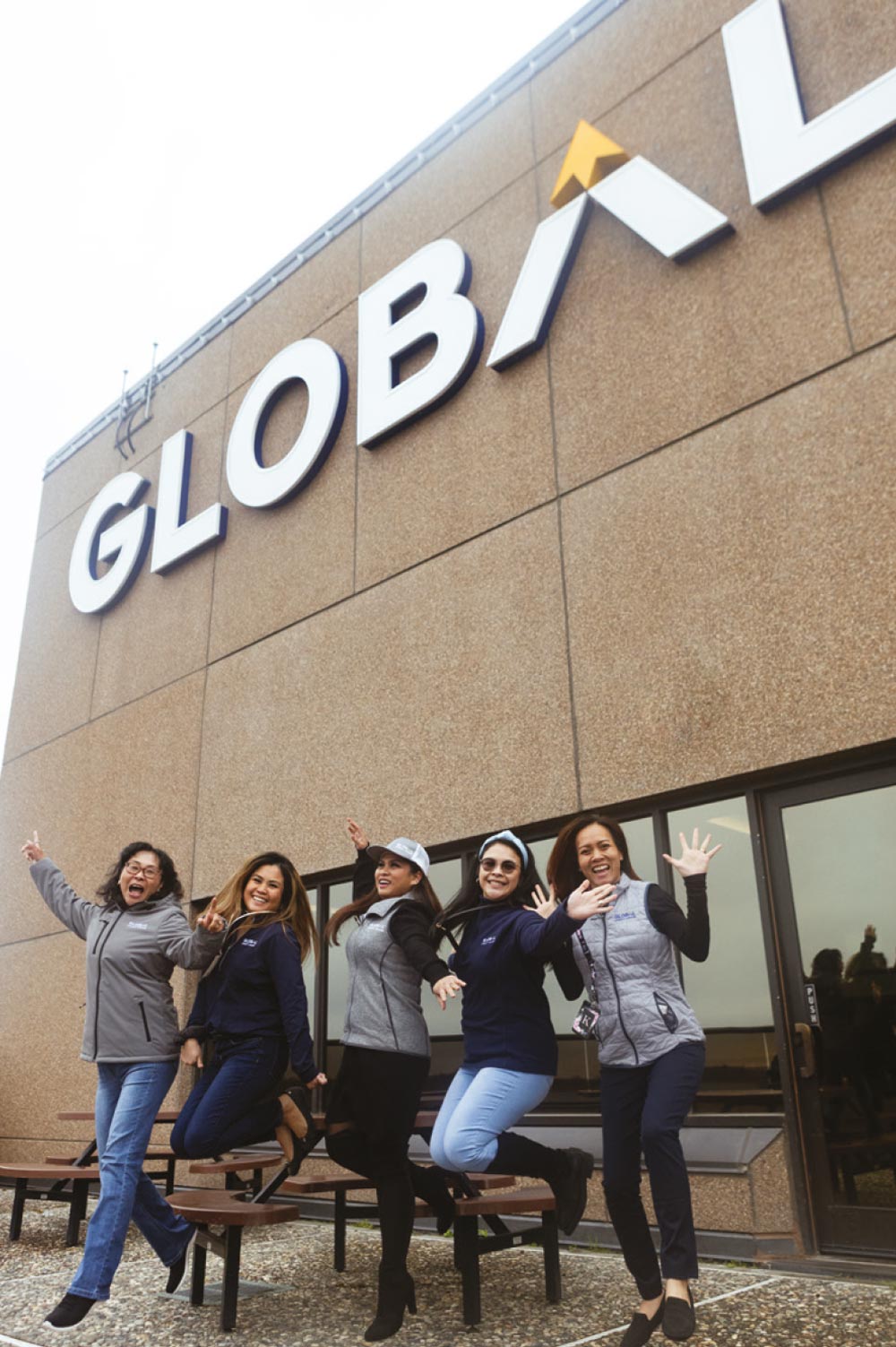Five women are jumping in unison in front of a building with a large "GLOBAL" sign on it.