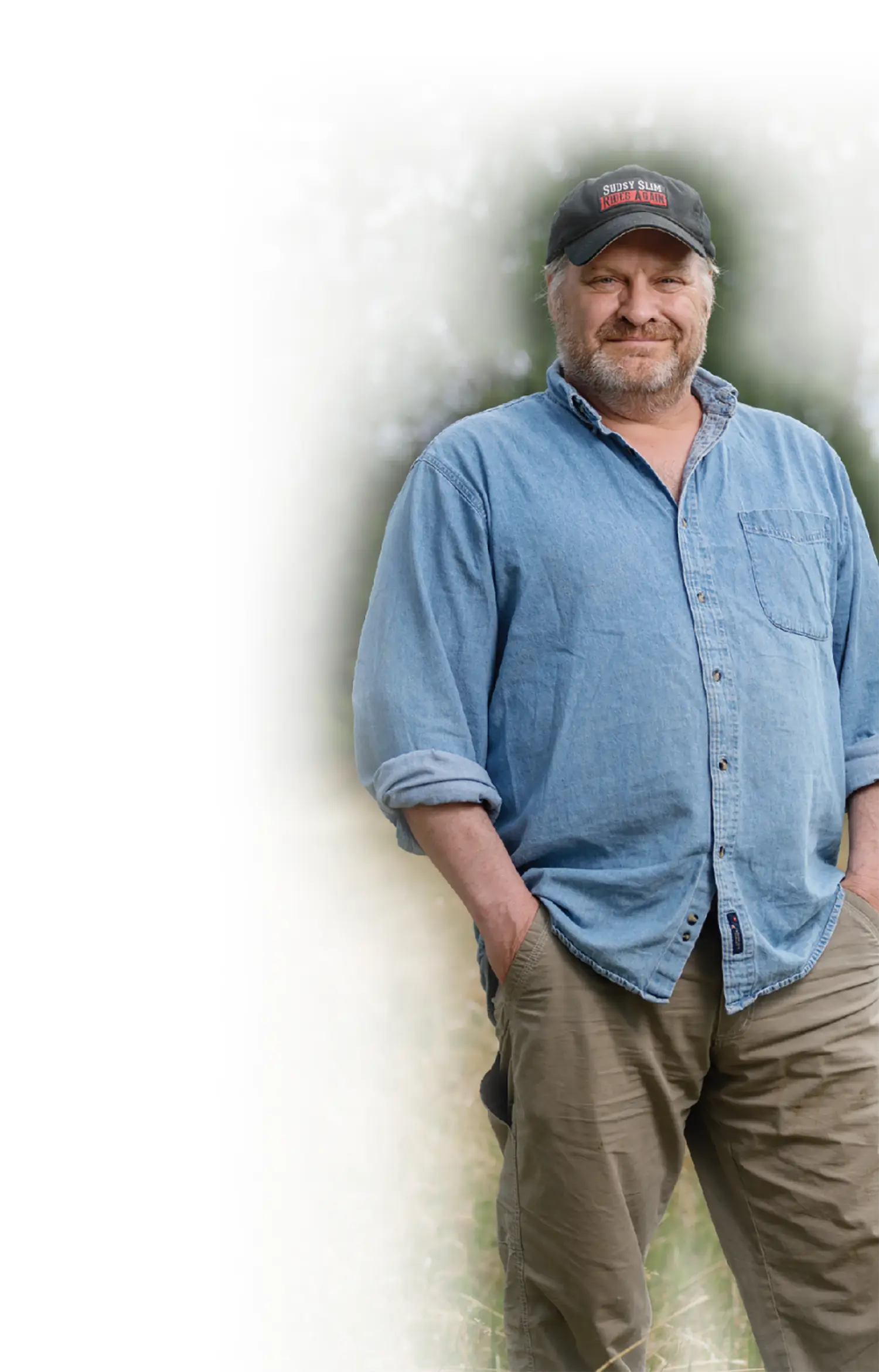 Mostly white to transparent gradient background covering the portrait orientation outdoor photograph close-up view of Chad Carpenter, a smiling man in a blue denim button-down dress shirt and dark faded charcoal black covered curved shaped hat that states the following phrase Sudsy Slim Rides Again as he has hands inside his dark brown khaki style pants with him standing outdoors in nature