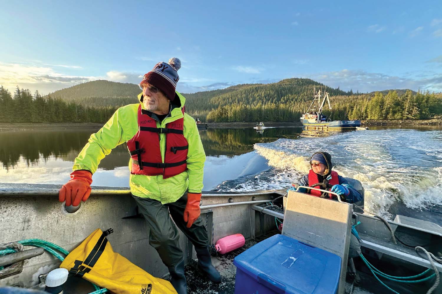 A person in a neon yellow jacket and red life vest stands in a boat, looking out at a forested shore under a bright sky. Another person is seated further back in the boat, near the steering. The boat is in motion, leaving a wake, and other boats are visible in the distance.