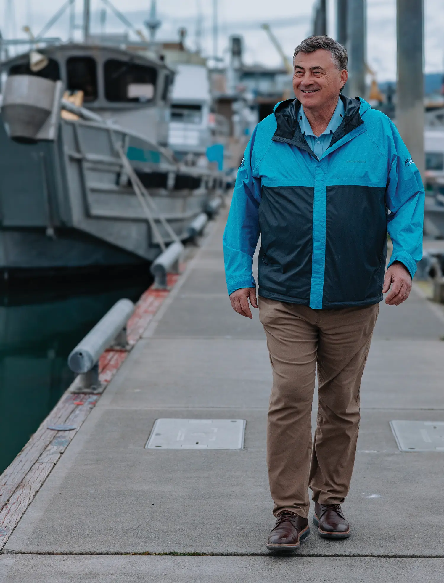 Portrait orientation outdoor photograph of John Hendrix, a smiling man wearing a GRUNDÉNS logo branded turquoise blue-colored and black jacket, a light turquoise blue-colored collared polo top dress shirt underneath, dark brown pants, and dark brown work dress shoes as he walks along on top of a concrete harbor dock pathway nearby a body of water; In the blurred background, various boats are docked and harbor structures are visible under an overcast sky; He is looking slightly to his right into the distance