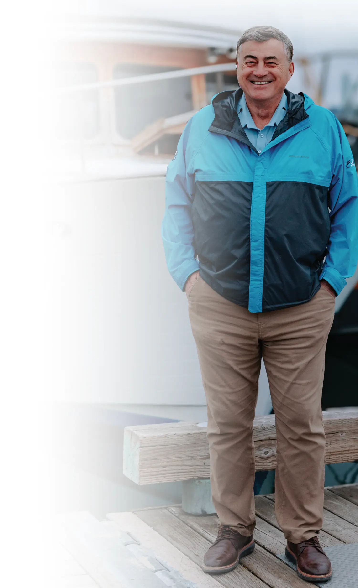 Mostly white gradient background covering the portrait orientation outdoor photograph of John Hendrix, a smiling man wearing a GRUNDÉNS logo branded turquoise blue-colored and black jacket, a light turquoise blue-colored collared polo top dress shirt underneath, dark brown pants, and dark brown work dress shoes as he is standing along on top of a concrete/wooden harbor dock pathway nearby a body of water with his hands inside his pockets; In the blurred background, there happens to be a boat docked within a harbor visible under an overcast sky; He is looking towards the direction of the camera