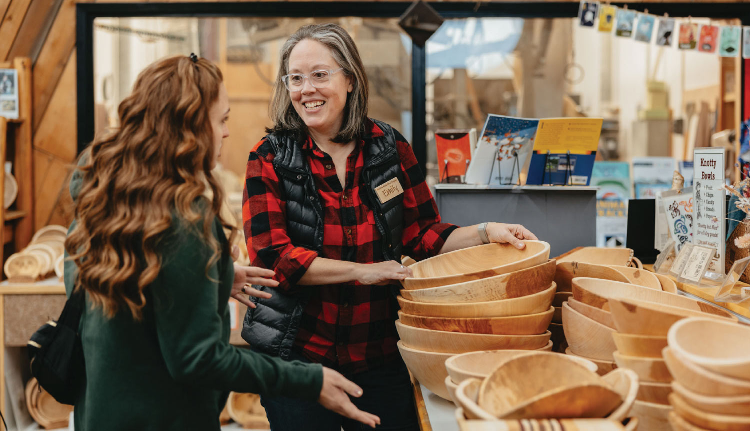 A medium, eye-level shot of two women talking in a store. The woman on the right is a sales clerk with a name tag that reads "Emily." She is smiling and gesturing with her hands towards a large display of wooden bowls. 