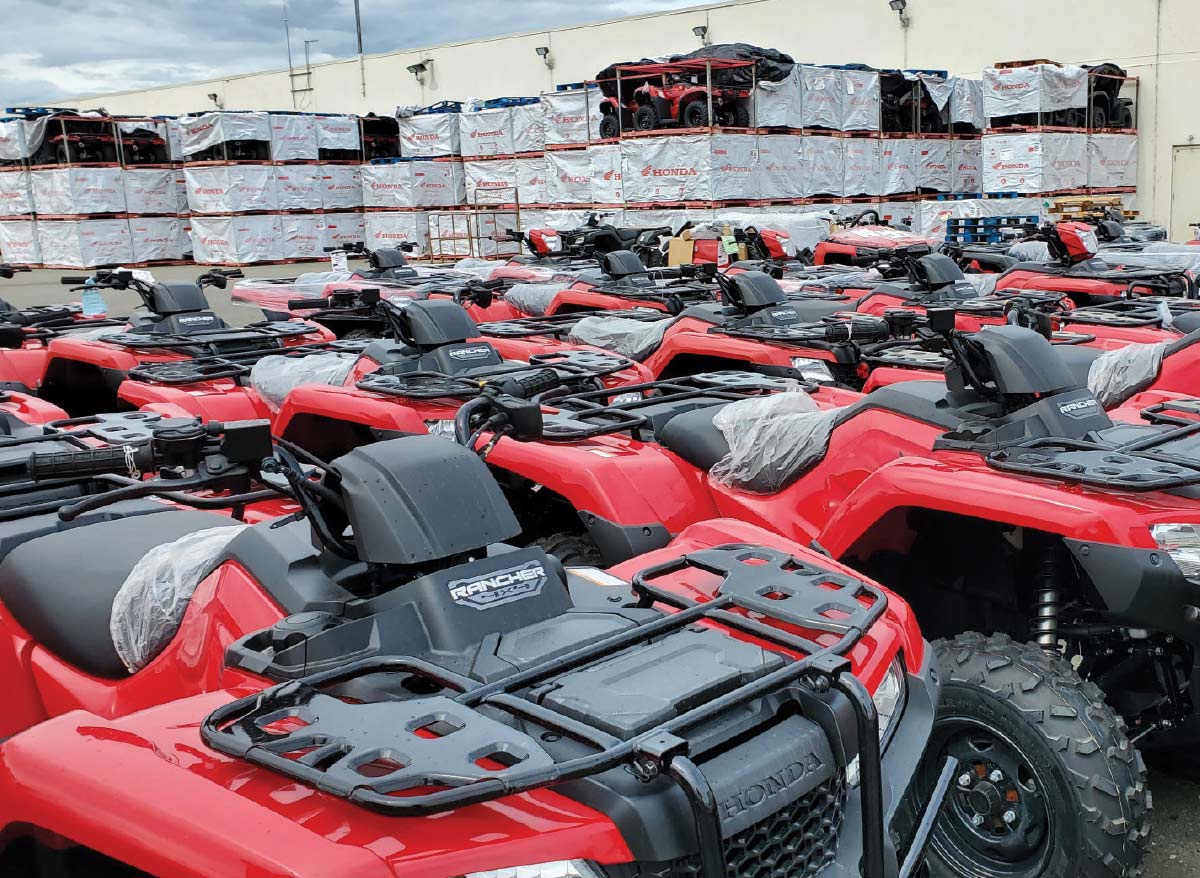 A front-facing medium shot of a large number of red Honda Rancher ATVs, parked outside in rows on a gray pavement. 
