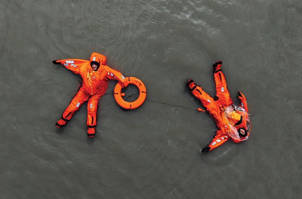Top view of two people in orange immersion suits floating in water, connected by a life ring.
