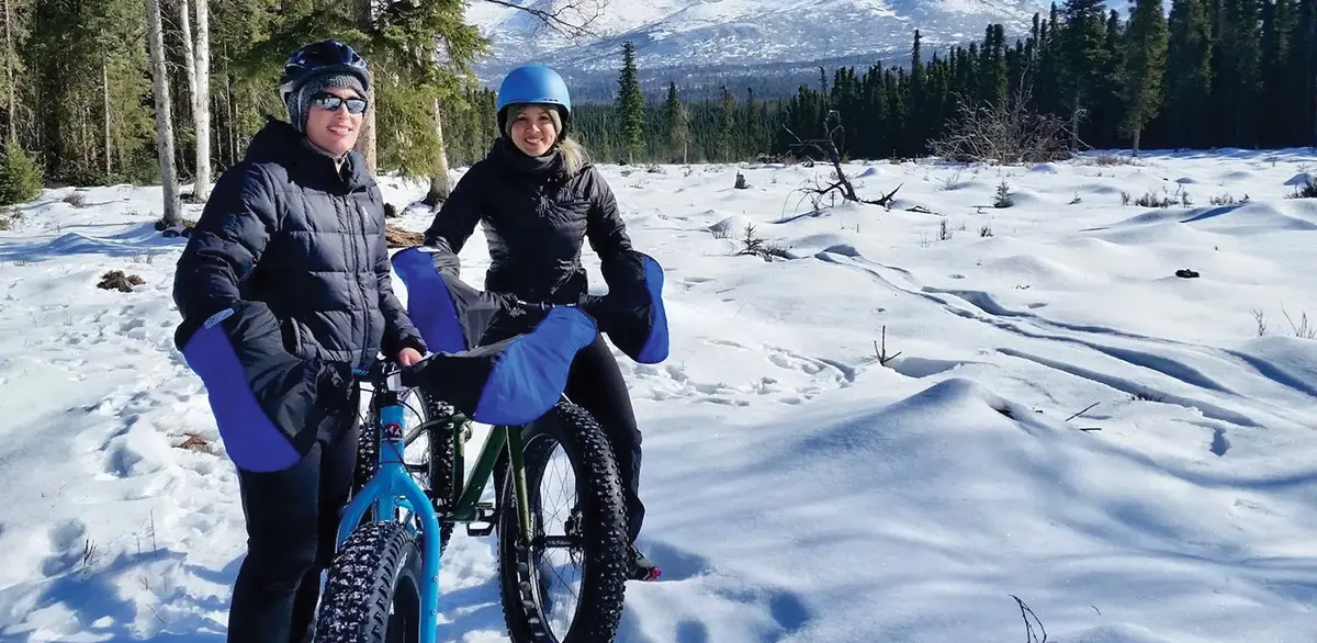 Two smiling bikers in winter gear standing in a snowy field with a mountain in the distance.