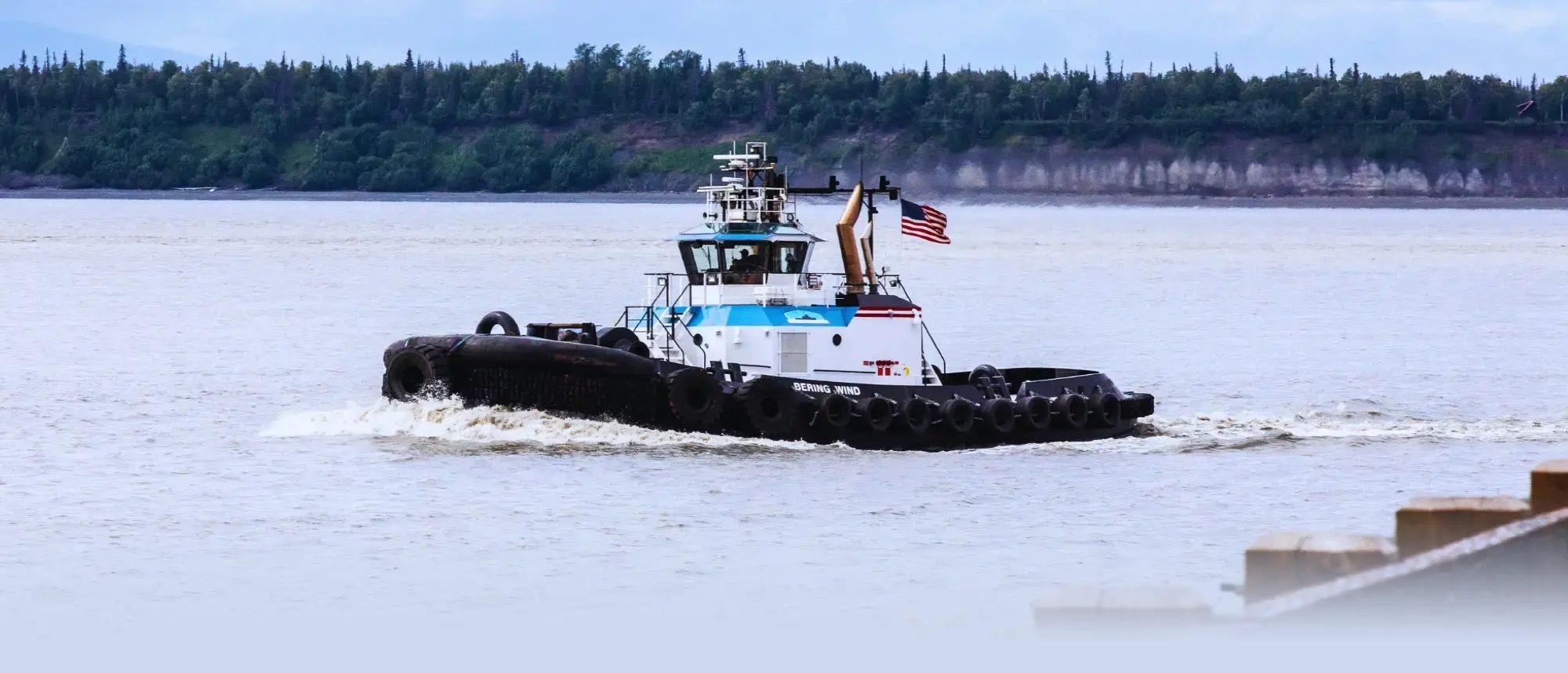 The Bering Wind tugboat on the water, with a lush green treeline and bluff behind it.